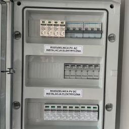 SKENERGY SPÓŁKA Z OGRANICZONĄ ODPOWIEDZIALNOŚCIĄ - Szara rozdzielnica PV AC/DC z otwartymi drzwiczkami, widoczne zabezpieczenia i naklejki ostrzegawcze. Instalacja elektryczna paneli fotowoltaicznych.