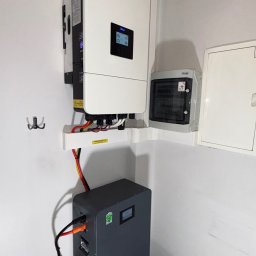 SKENERGY SPÓŁKA Z OGRANICZONĄ ODPOWIEDZIALNOŚCIĄ - Zainstalowany biały inwerter fotowoltaiczny firmy Days z szarą baterią magazynu energii Eco ISO w rogu pomieszczenia z szarą podłogą i rurą wentylacyjną.