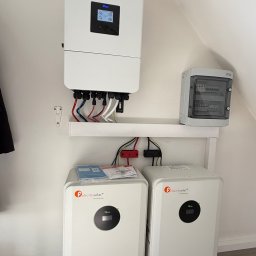 SKENERGY SPÓŁKA Z OGRANICZONĄ ODPOWIEDZIALNOŚCIĄ - Instalacja magazynu energii Felicity Solar z inwerterem Deye i skrzynką bezpiecznikową na białej ścianie. Widoczne kable i dokumentacja.