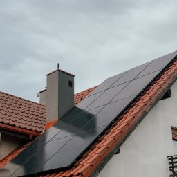 SKENERGY SPÓŁKA Z OGRANICZONĄ ODPOWIEDZIALNOŚCIĄ - Montaż paneli fotowoltaicznych na dachu domu jednorodzinnego z czerwoną dachówką. Widoczny monter na drabinie, instalacja elektryczna i fragment elewacji.