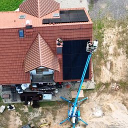 SKENERGY SPÓŁKA Z OGRANICZONĄ ODPOWIEDZIALNOŚCIĄ - Instalacja paneli słonecznych na dachu domu z perspektywy lotniczej. Na ziemi podnośnik koszowy, mini koparka i samochód dostawczy. Prace w toku.