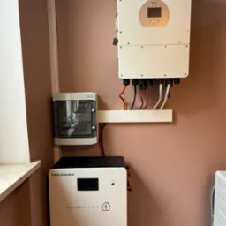 Kompletna instalacja: magazyn energii Heckman, inwerter Deye i rozdzielnica.  Widoczne przewody i elementy montażowe. Realizacja w pomieszczeniu o beżowych ścianach.