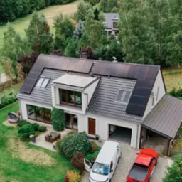 Dom z panelami słonecznymi na dachu, widok z góry. Na podjeździe biały van z otwartymi drzwiami i czerwony pickup. W tle zalesione wzgórza.