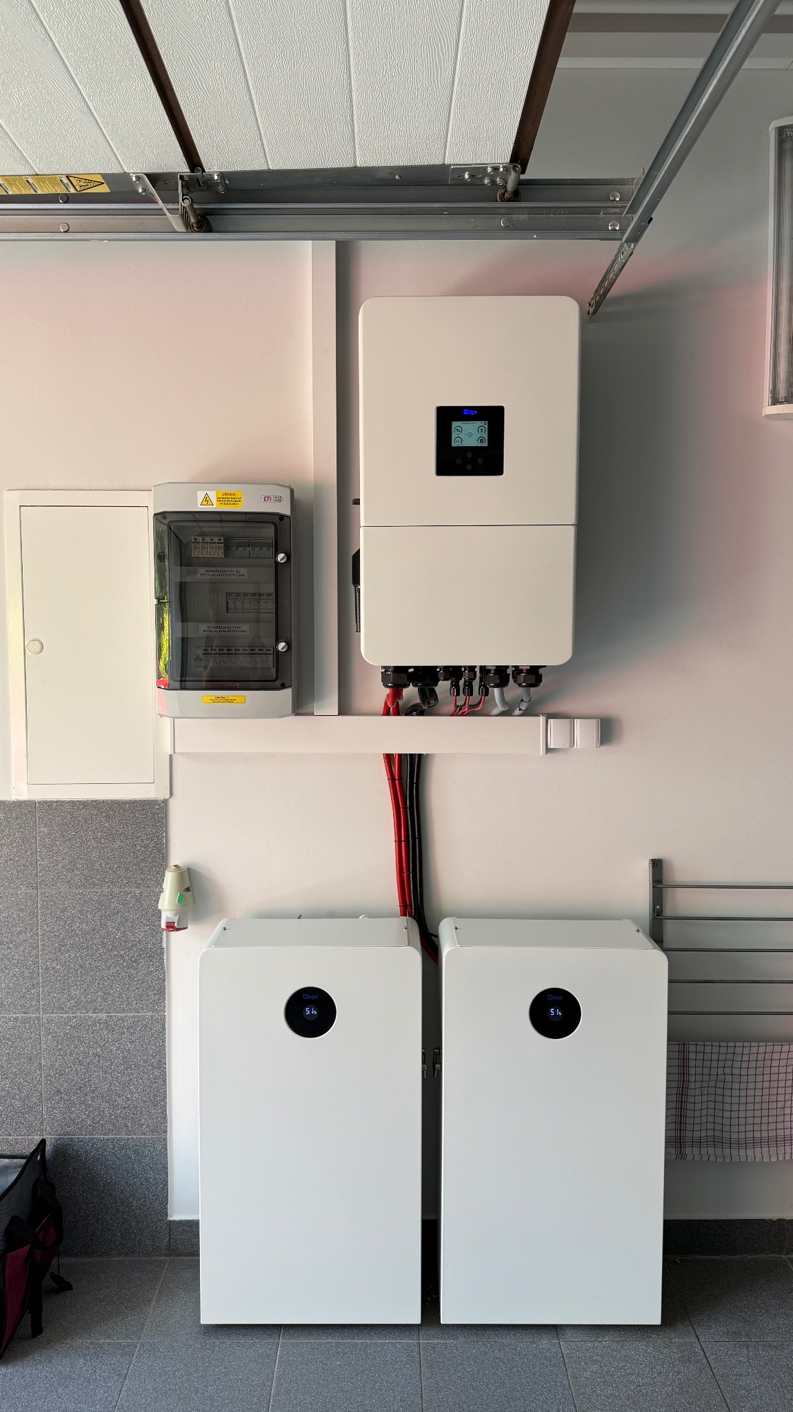 Białe magazyny energii z inwerterem na ścianie garażu. Widoczna skrzynka elektryczna i instalacja. Szara podłoga i brama garażowa w tle.