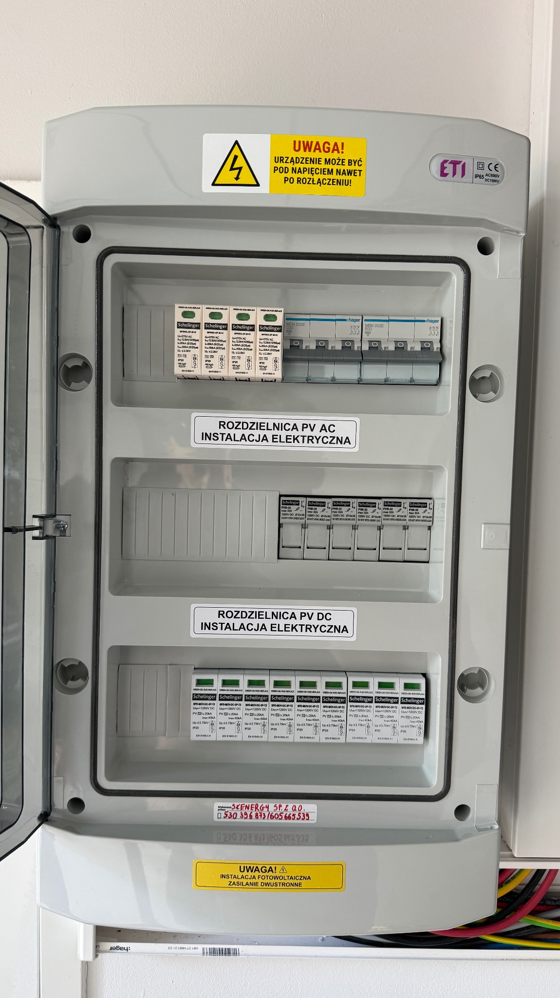 Szara rozdzielnica PV AC/DC z otwartymi drzwiczkami, widoczne zabezpieczenia i naklejki ostrzegawcze. Instalacja elektryczna paneli fotowoltaicznych.