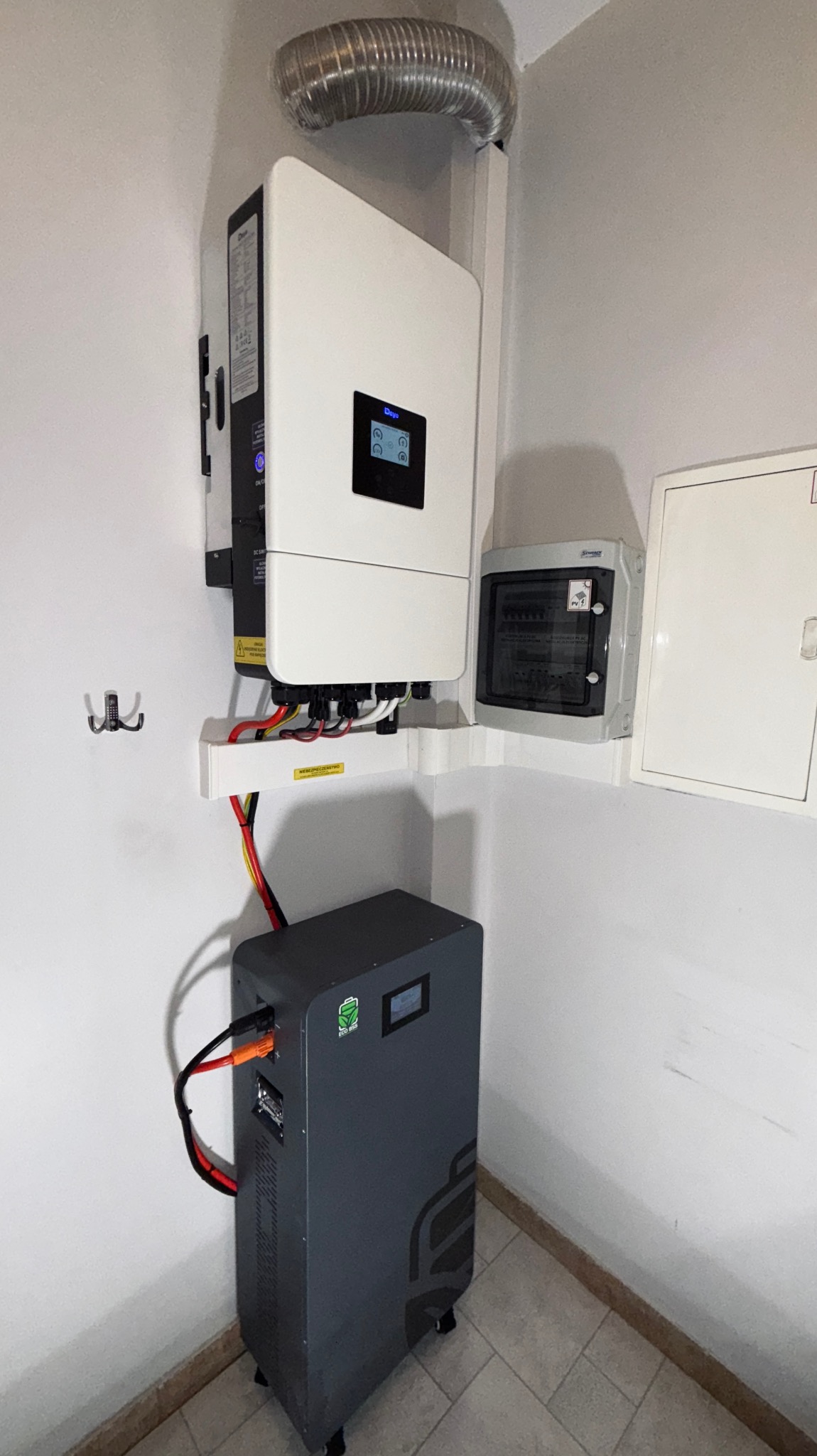 Zainstalowany biały inwerter fotowoltaiczny firmy Days z szarą baterią magazynu energii Eco ISO w rogu pomieszczenia z szarą podłogą i rurą wentylacyjną.