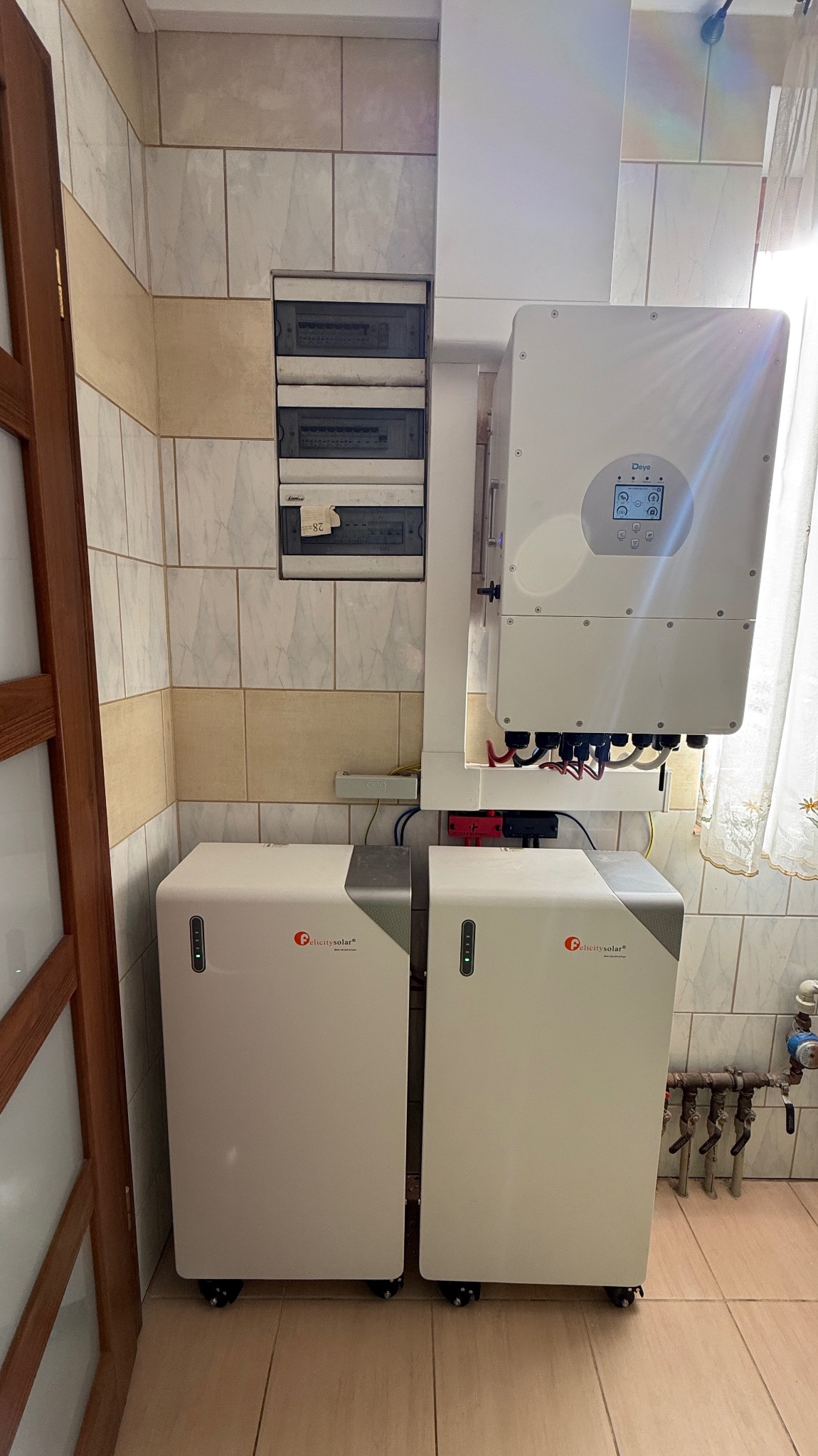 Domowa instalacja: dwa magazyny energii Felicitysolar obok inwertera Deye, z widoczną skrzynką bezpiecznikową na ścianie z beżowych płytek. Estetyczne rozwiązanie.