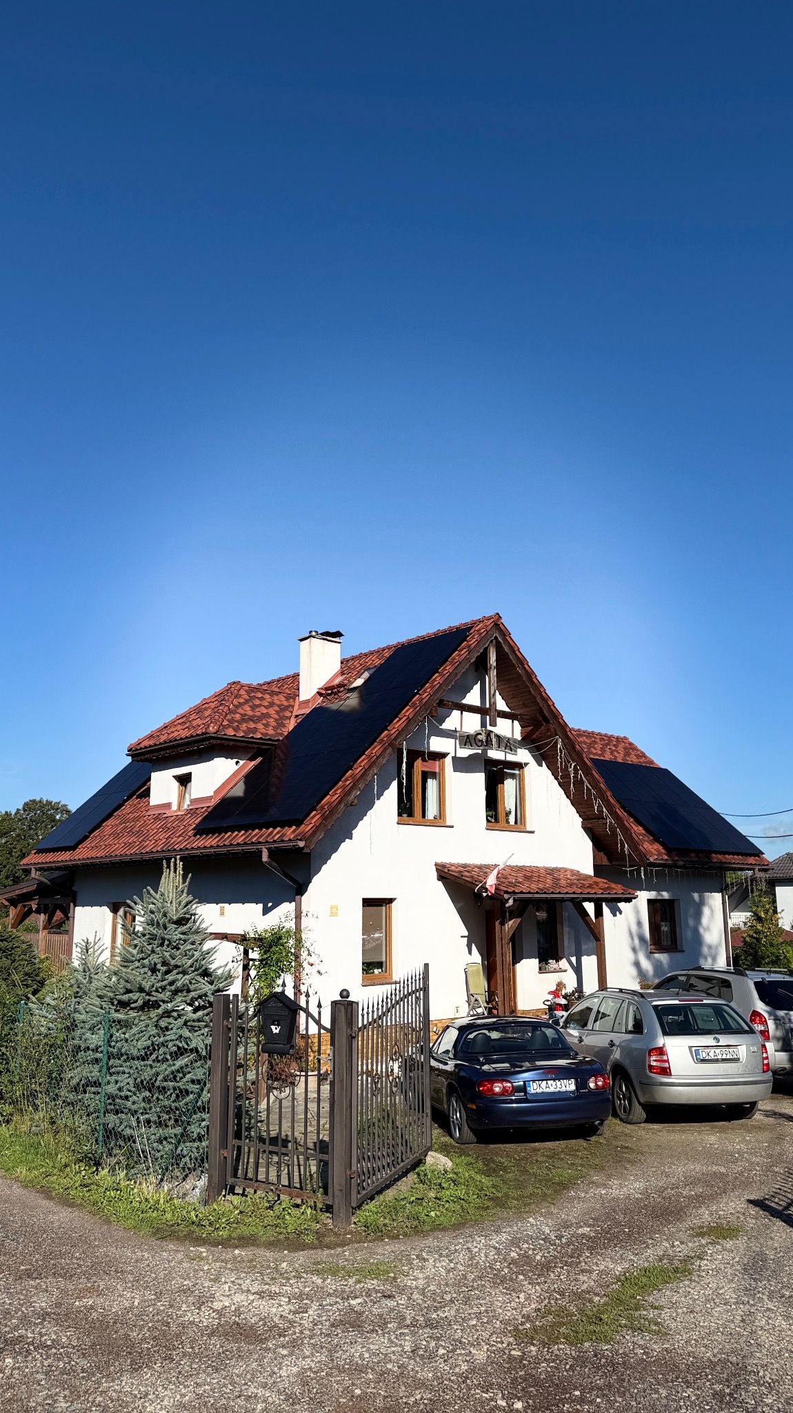 Biały dom z czerwoną dachówką i panelami słonecznymi. Na podjeździe zaparkowane samochody. Błękitne niebo w tle.