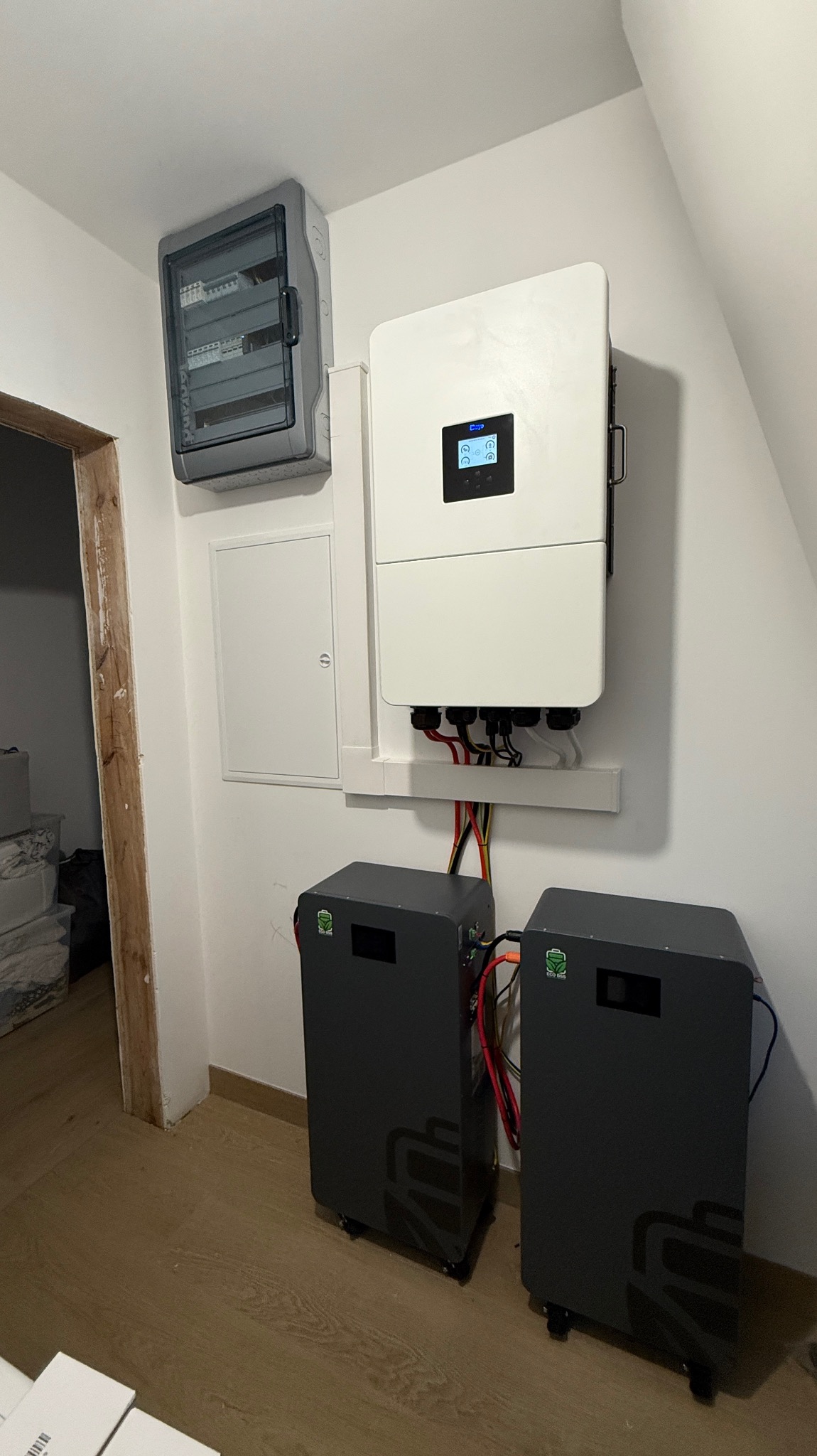 Kompletna instalacja fotowoltaiczna w pomieszczeniu: inwerter, dwa akumulatory energii z logotypem i rozdzielnica elektryczna na białej ścianie.