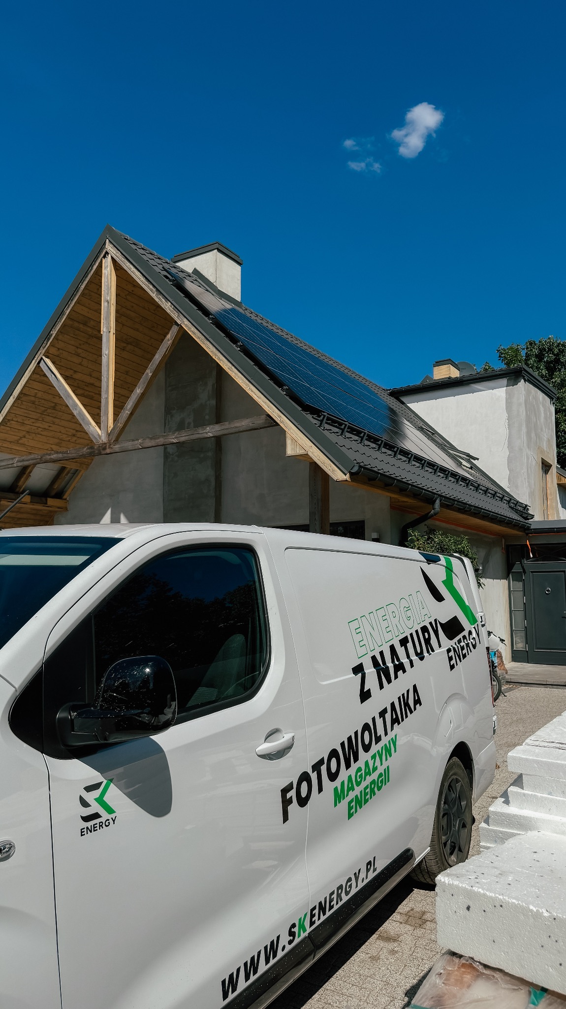 Instalacja paneli słonecznych na dachu domu, widoczna zza firmowego samochodu z logo i napisem 'Fotowoltaika Magazyny Energii'. Błękitne niebo w tle.