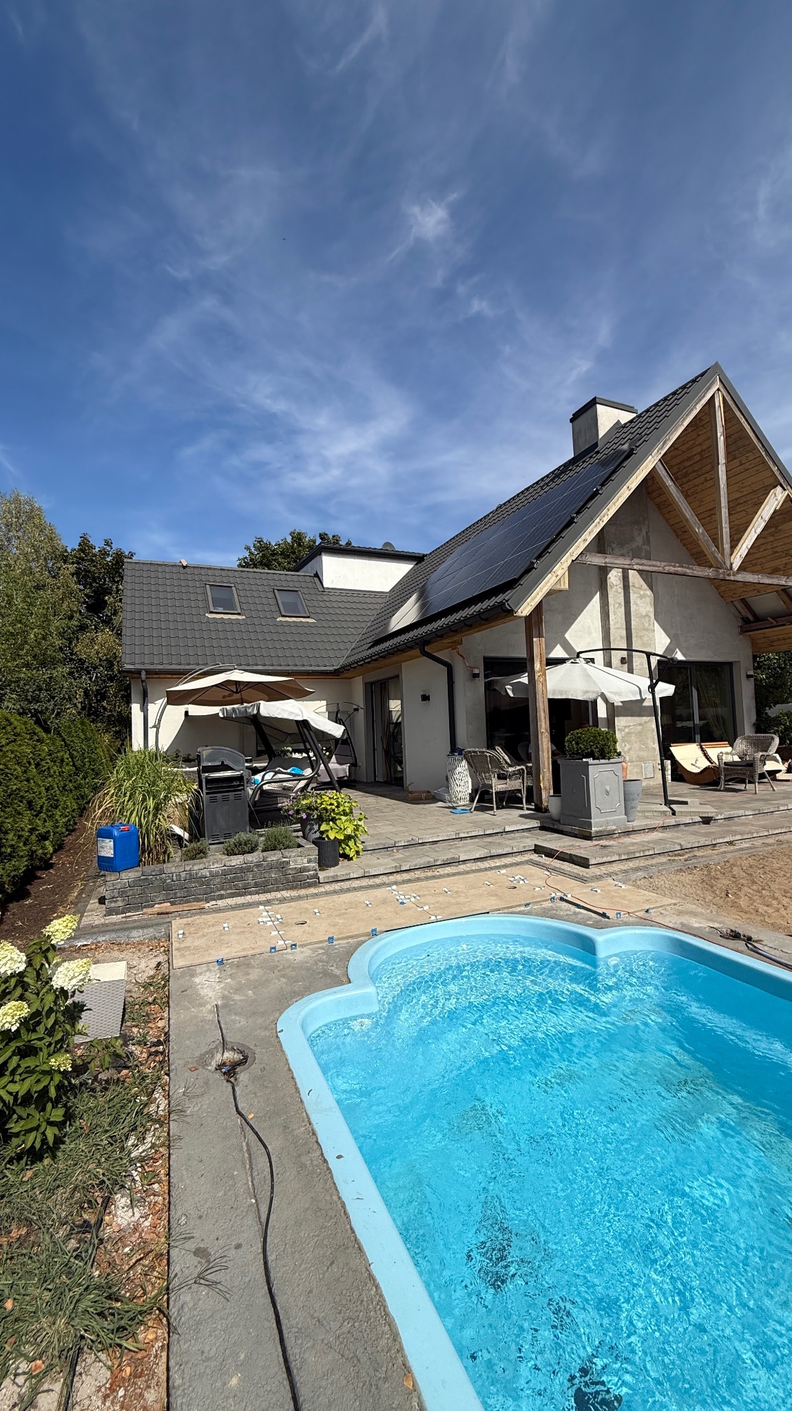 Dom jednorodzinny z panelami słonecznymi na dachu, widok z perspektywy basenu. Na tarasie meble ogrodowe i parasole. Błękitne niebo z delikatnymi chmurami w tle.