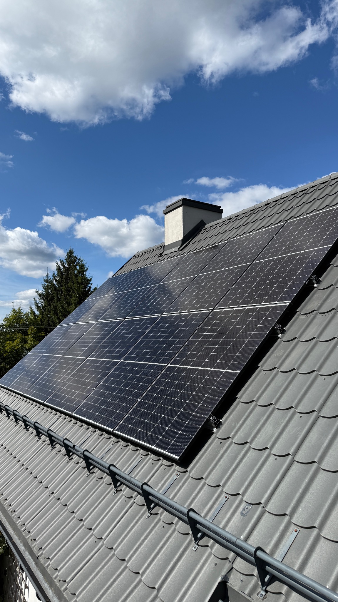 Panele fotowoltaiczne na szarym dachu z dachówki ceramicznej, widoczne mocowania, w tle błękitne niebo z chmurami i komin. Widok z dołu.