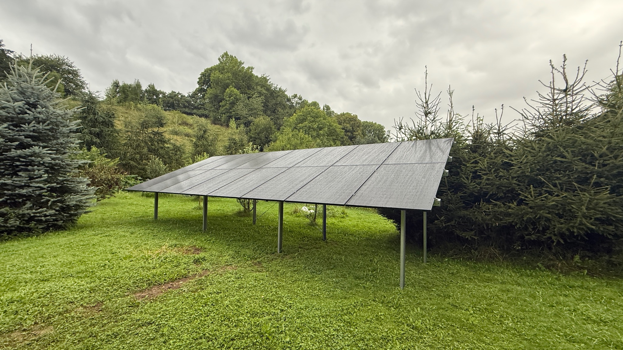 Panel słoneczny na metalowej konstrukcji, otoczony zielenią i drzewami, pod pochmurnym niebem. Widok z perspektywy lekko z dołu, podkreślający konstrukcję paneli.