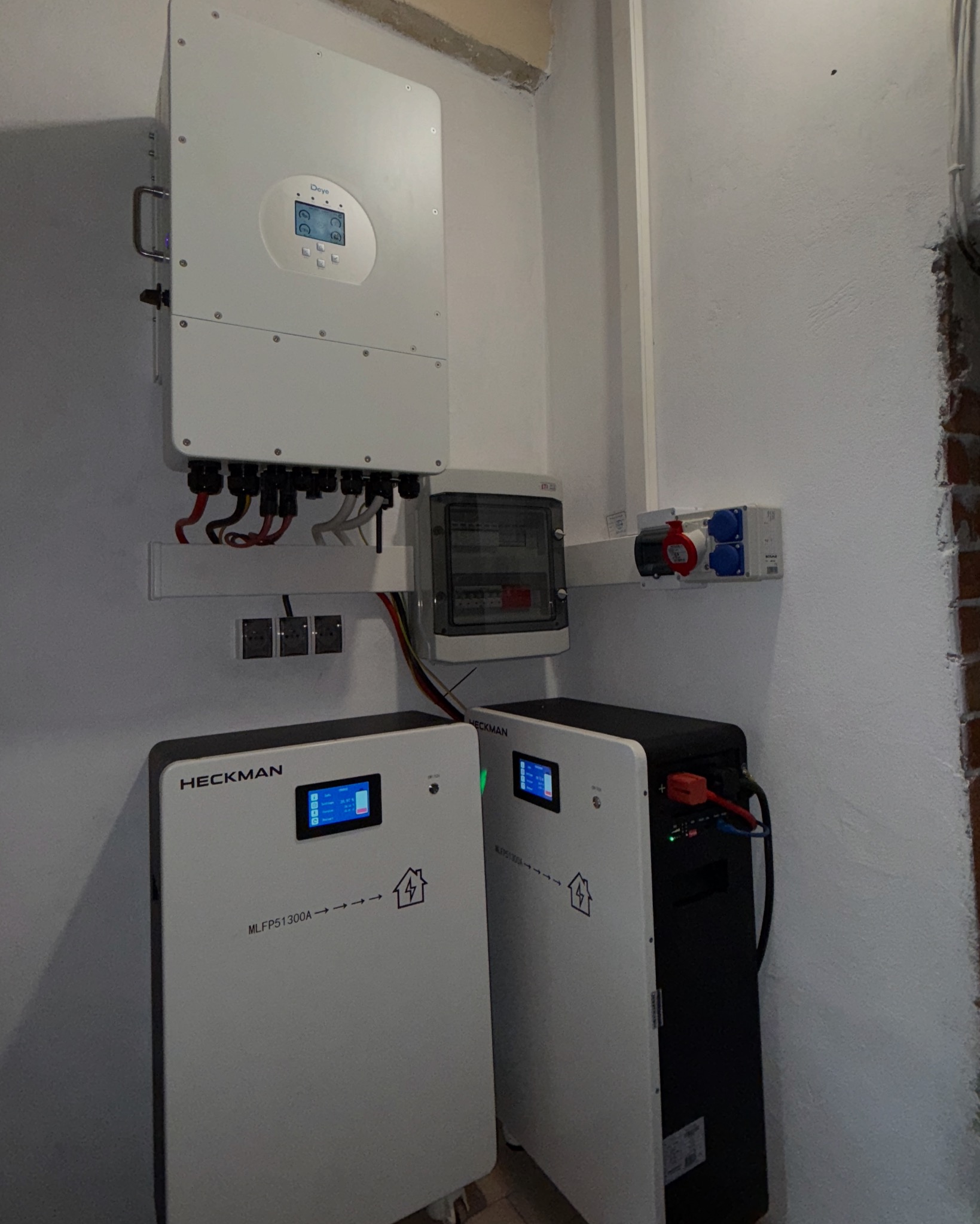 Instalacja magazynu energii Heckman MLFP51300A z inwerterem Deye, z widoczną rozdzielnicą i gniazdami, zamontowana na białej ścianie w pomieszczeniu.