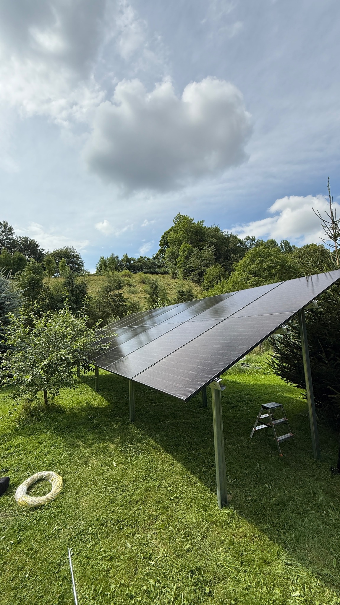 Instalacja paneli słonecznych na trawie w Kamiennej Górze. Widoczne panele, drabina, zwój kabla i okoliczna zieleń. Słoneczny dzień, lekkie zachmurzenie.