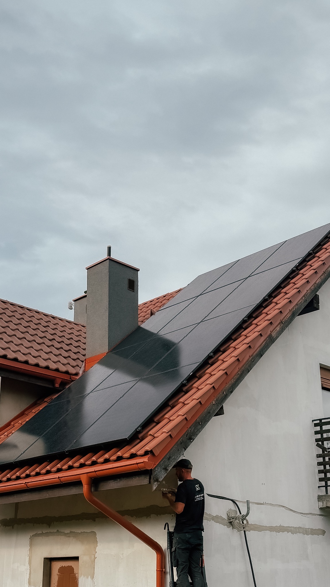 Montaż paneli fotowoltaicznych na dachu domu jednorodzinnego z czerwoną dachówką. Widoczny monter na drabinie, instalacja elektryczna i fragment elewacji.