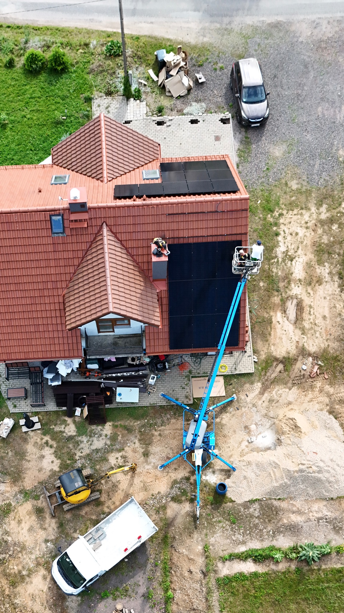 Instalacja paneli słonecznych na dachu domu z perspektywy lotniczej. Na ziemi podnośnik koszowy, mini koparka i samochód dostawczy. Prace w toku.