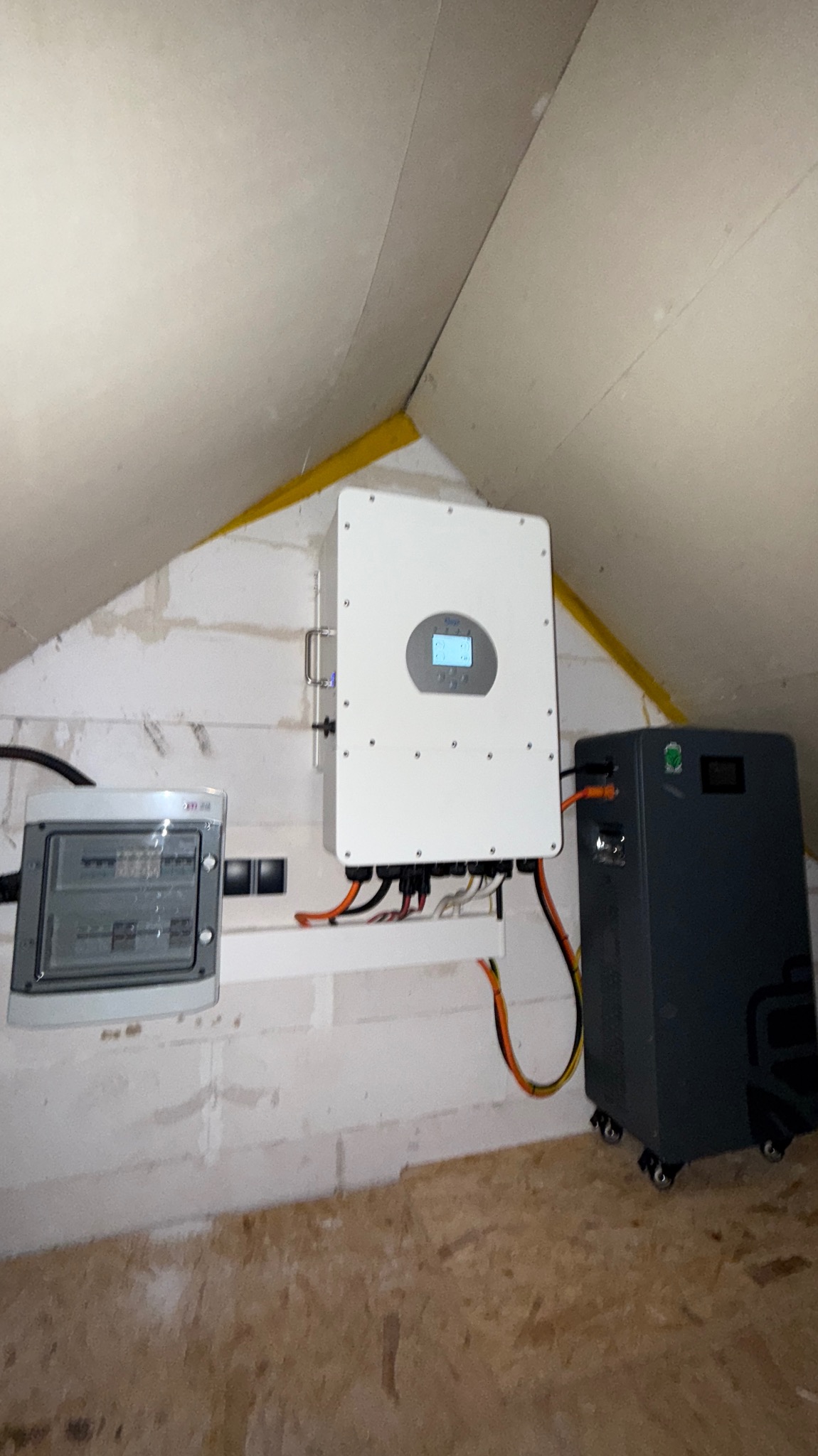 Instalacja: biały inwerter, szara skrzynka rozdzielcza i akumulator na kółkach w pomieszczeniu ze skośnym sufitem, widoczne przewody.