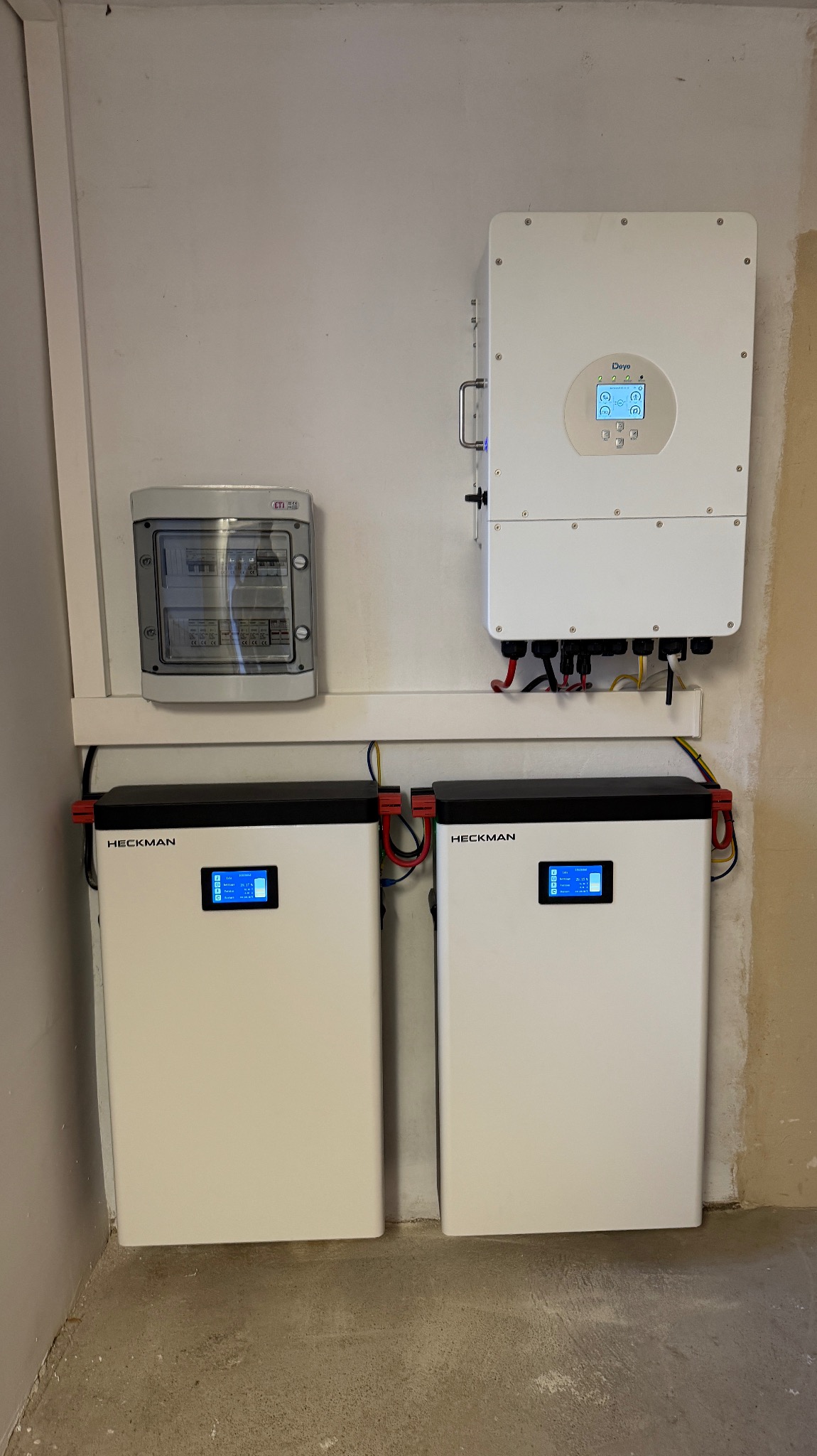 Instalacja domowa: dwa białe magazyny energii Heckman, inwerter Deye i skrzynka z bezpiecznikami na białej ścianie. Widoczne kable i przewody.