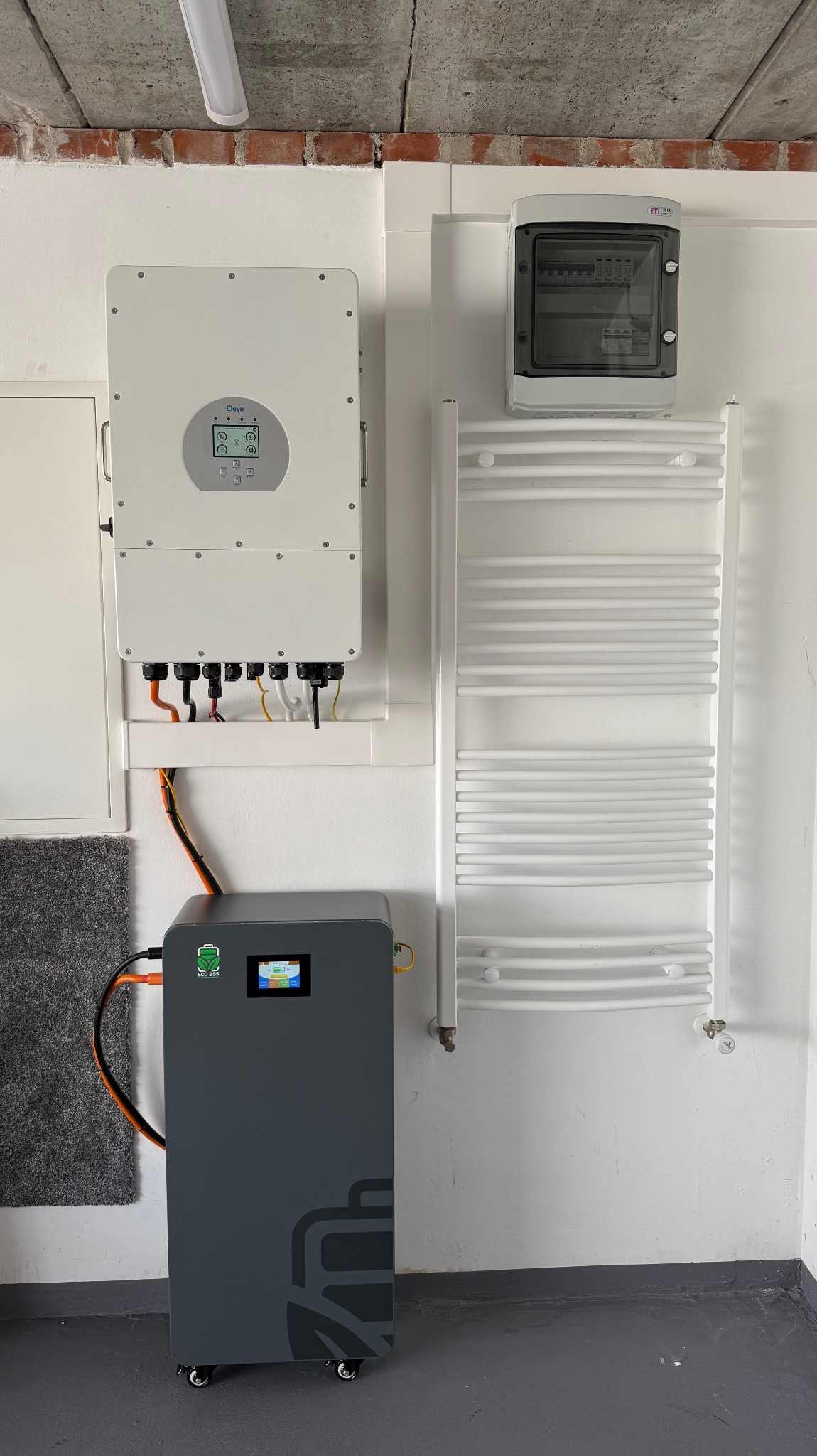 Szary magazyn energii ECO BSS na kółkach, podłączony do białego inwertera DEYE na ścianie, z widoczną skrzynką bezpiecznikową i grzejnikiem.