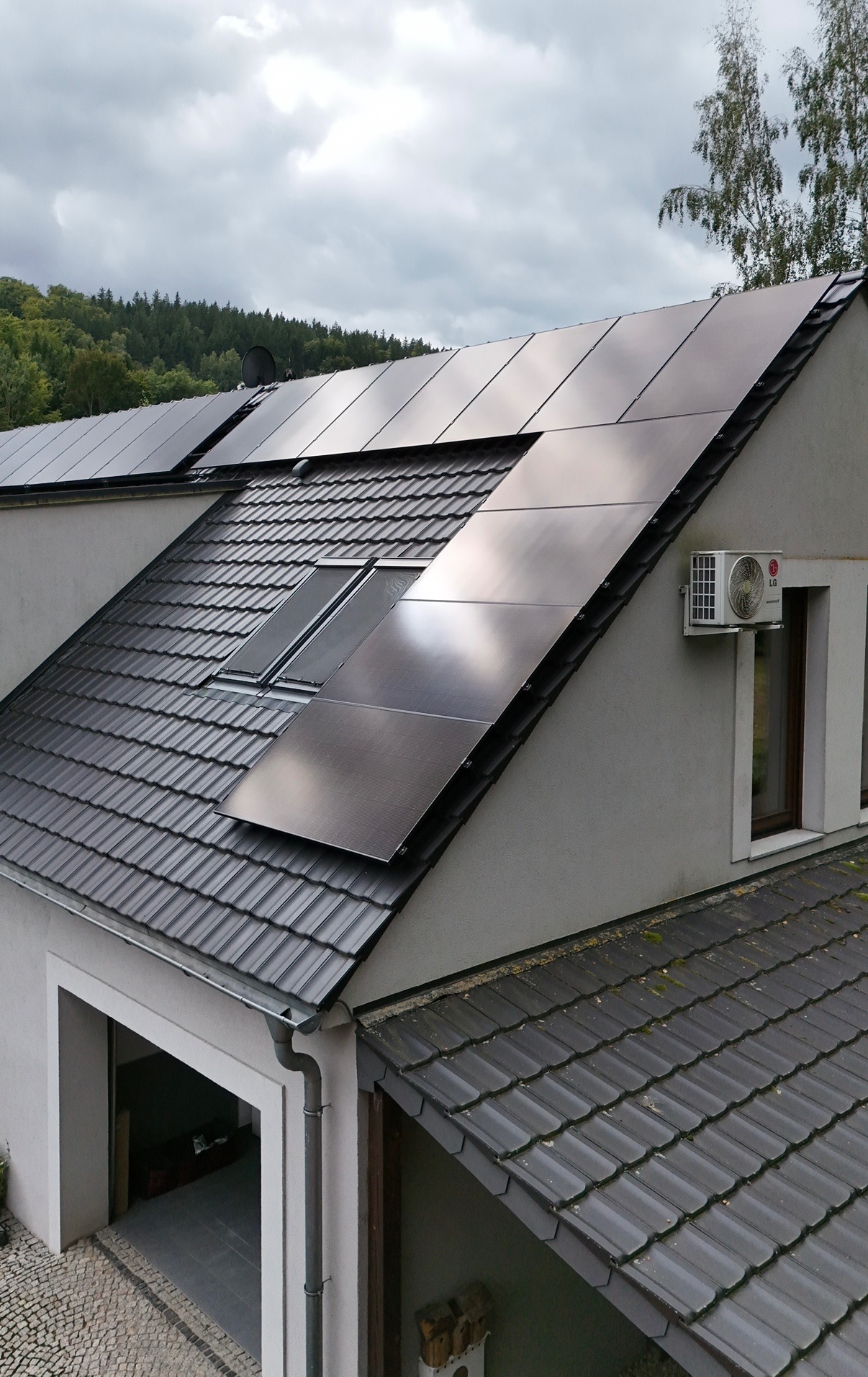 Instalacja fotowoltaiczna na dachu z dachówką ceramiczną, widok z góry. Na ścianie zamontowany klimatyzator. Widoczne okna dachowe i elementy orynnowania.