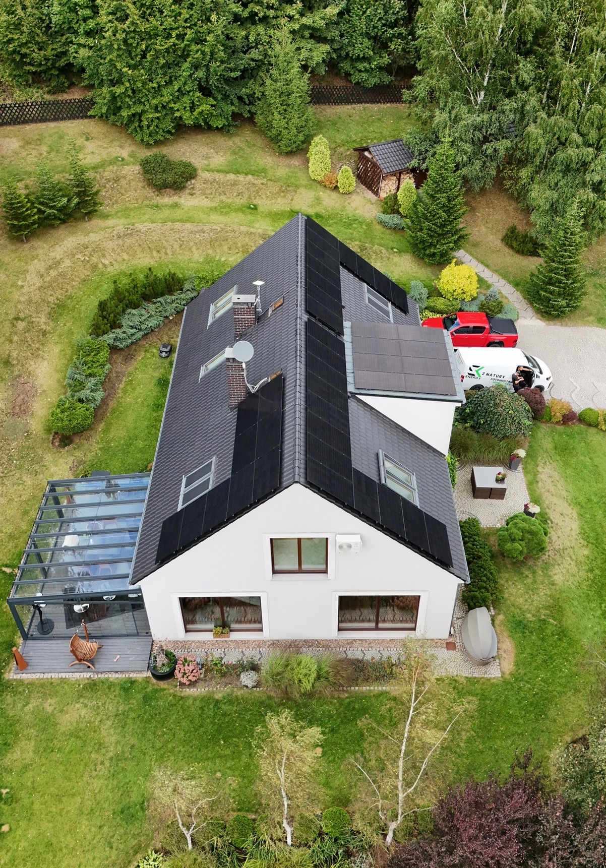 Dom z panelami słonecznymi na dachu, widziany z góry. Na podjeździe stoi firmowy samochód z logo instalatora. Otoczenie domu to zadbany ogród.
