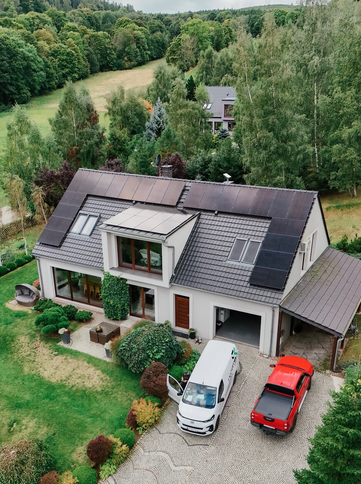 Dom z panelami słonecznymi na dachu, widok z góry. Na podjeździe biały van z otwartymi drzwiami i czerwony pickup. W tle zalesione wzgórza.