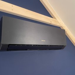 FL-System Grzegorz Górski - Czarny klimatyzator marki Gree zamontowany na granatowej ścianie pod drewnianą konstrukcją dachu.