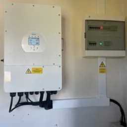 Biały inwerter Deye i rozdzielnica PV-AC na ścianie. Widoczne kable w peszlach. Żółte naklejki ostrzegawcze o napięciu. Montaż instalacji fotowoltaicznej wewnątrz budynku.