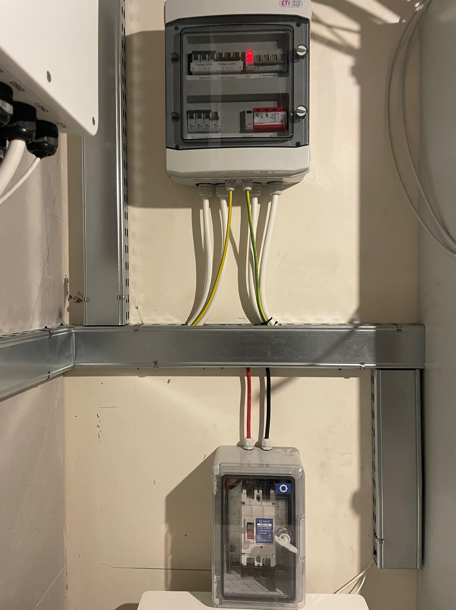 Rozdzielnica elektryczna z zabezpieczeniami, zamontowana na ścianie, z widocznymi przewodami w peszlach i metalowych korytkach kablowych.