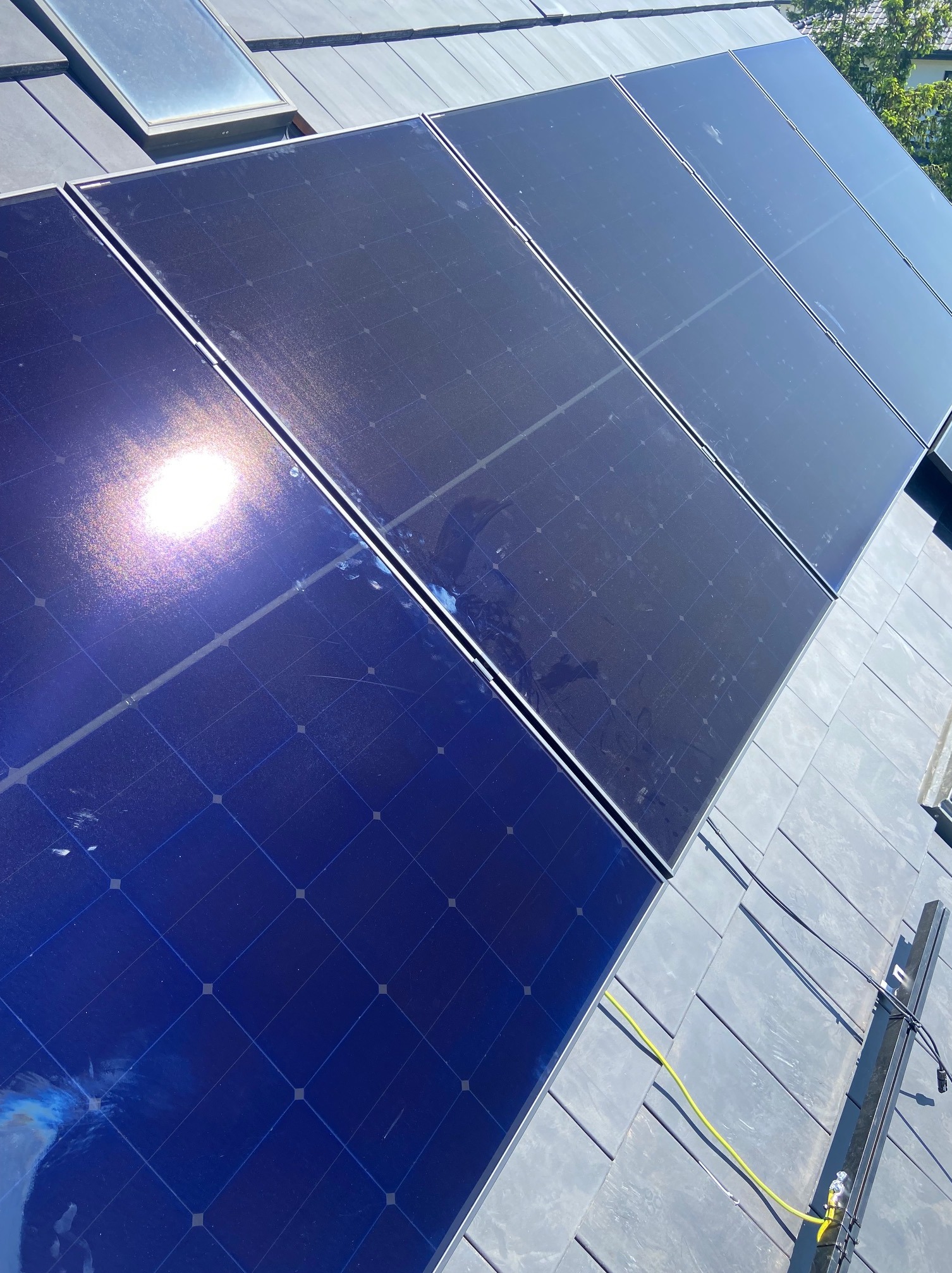 Panele słoneczne zamontowane na grafitowym dachu z widocznym świetlikiem, odblask słońca na jednym z paneli, żółty przewód i element konstrukcyjny widoczne w prawym dolnym rogu.