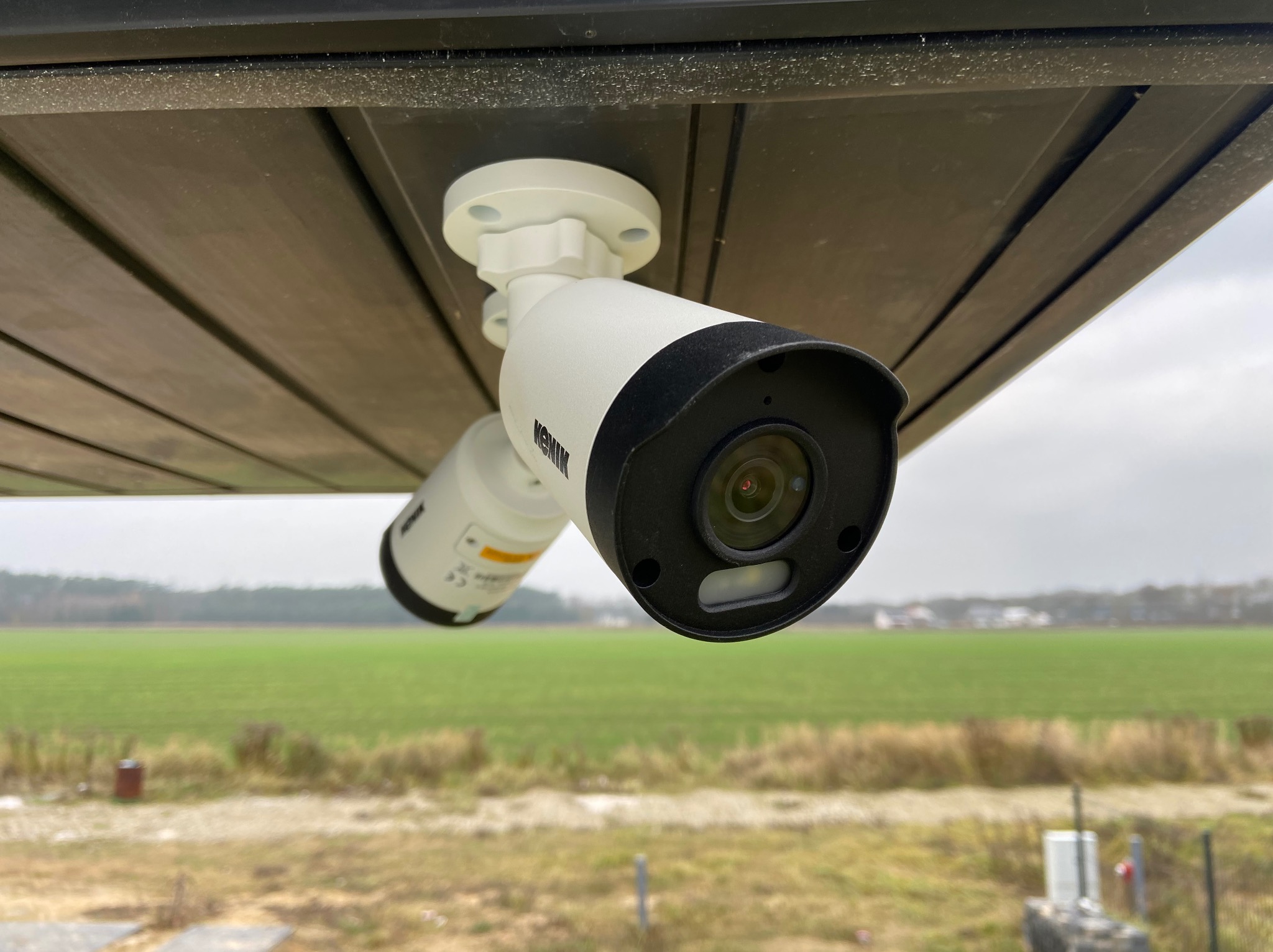 Dwie kamery monitoringu marki Kenik zamontowane pod zadaszeniem, z widokiem na zielone pole w Bobolinie. Widoczny obiektyw kamery z czarną obudową oraz biała podstawa mocująca.