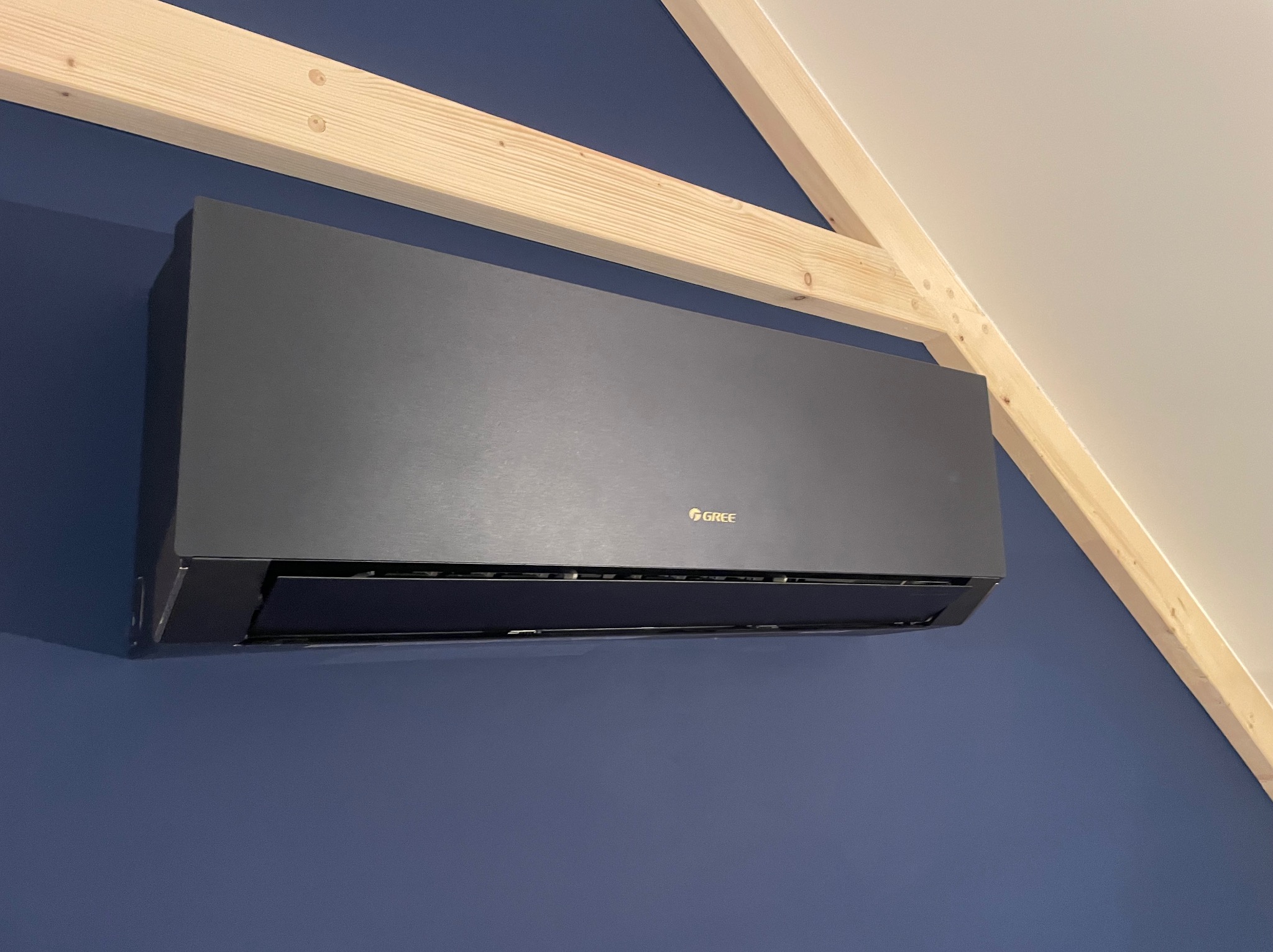 Czarny klimatyzator marki Gree zamontowany na granatowej ścianie pod drewnianą konstrukcją dachu.