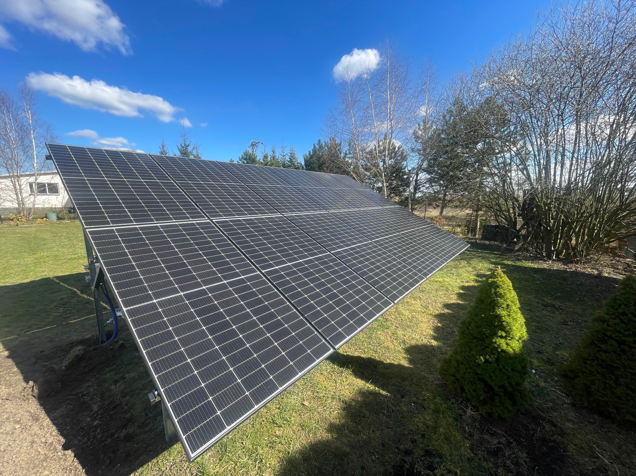 Panel słoneczny na trawniku w słoneczny dzień, widoczne niebieskie przewody i świeżo obsypana ziemia wokół konstrukcji.