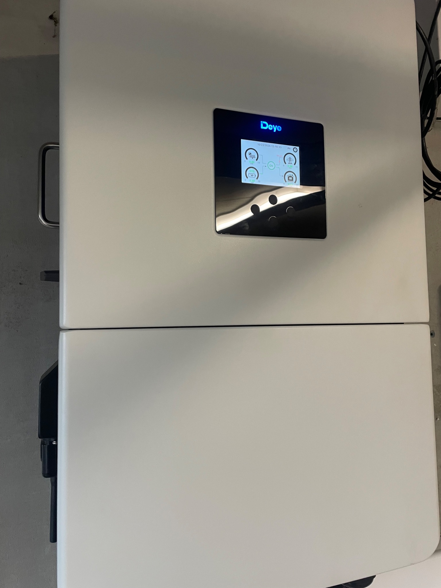 Biały magazyn energii Deye z czarnym panelem sterowania, wyświetlającym parametry pracy. Widoczne kable i uchwyt. Zdjęcie z bliska, lekko od dołu.