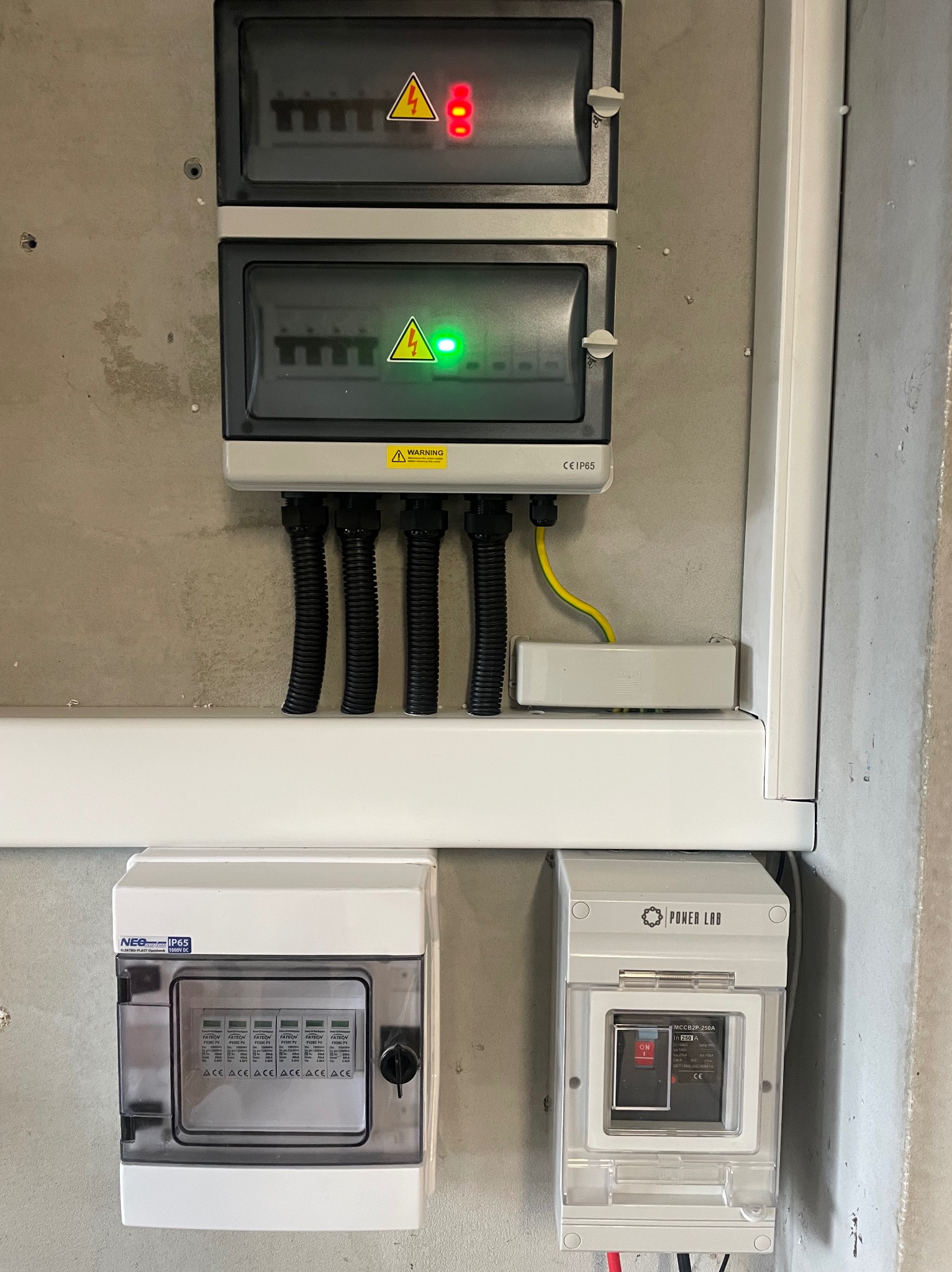 Rozdzielnia elektryczna z zabezpieczeniami, widoczne kable w peszlach,  lampki kontrolne (czerwona i zielona) oraz aparatura modułowa.  Szara ściana w tle.