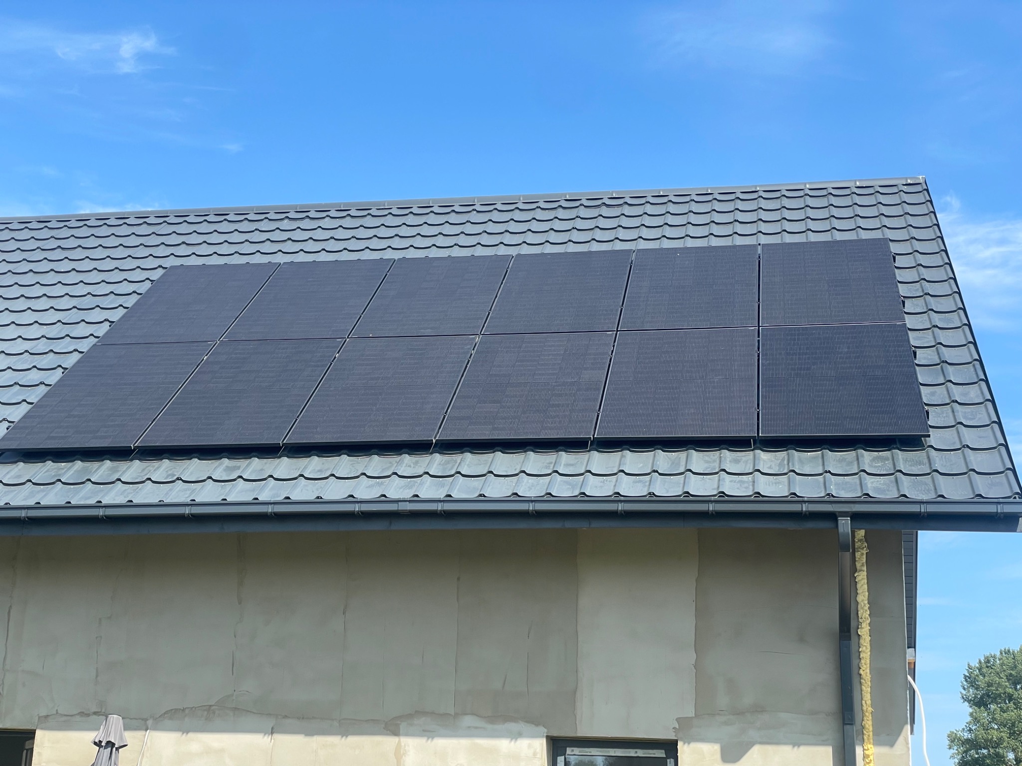 Panele fotowoltaiczne na dachu domu z szarej blachodachówki w słoneczny dzień. Widoczny fragment elewacji budynku w trakcie wykończenia.