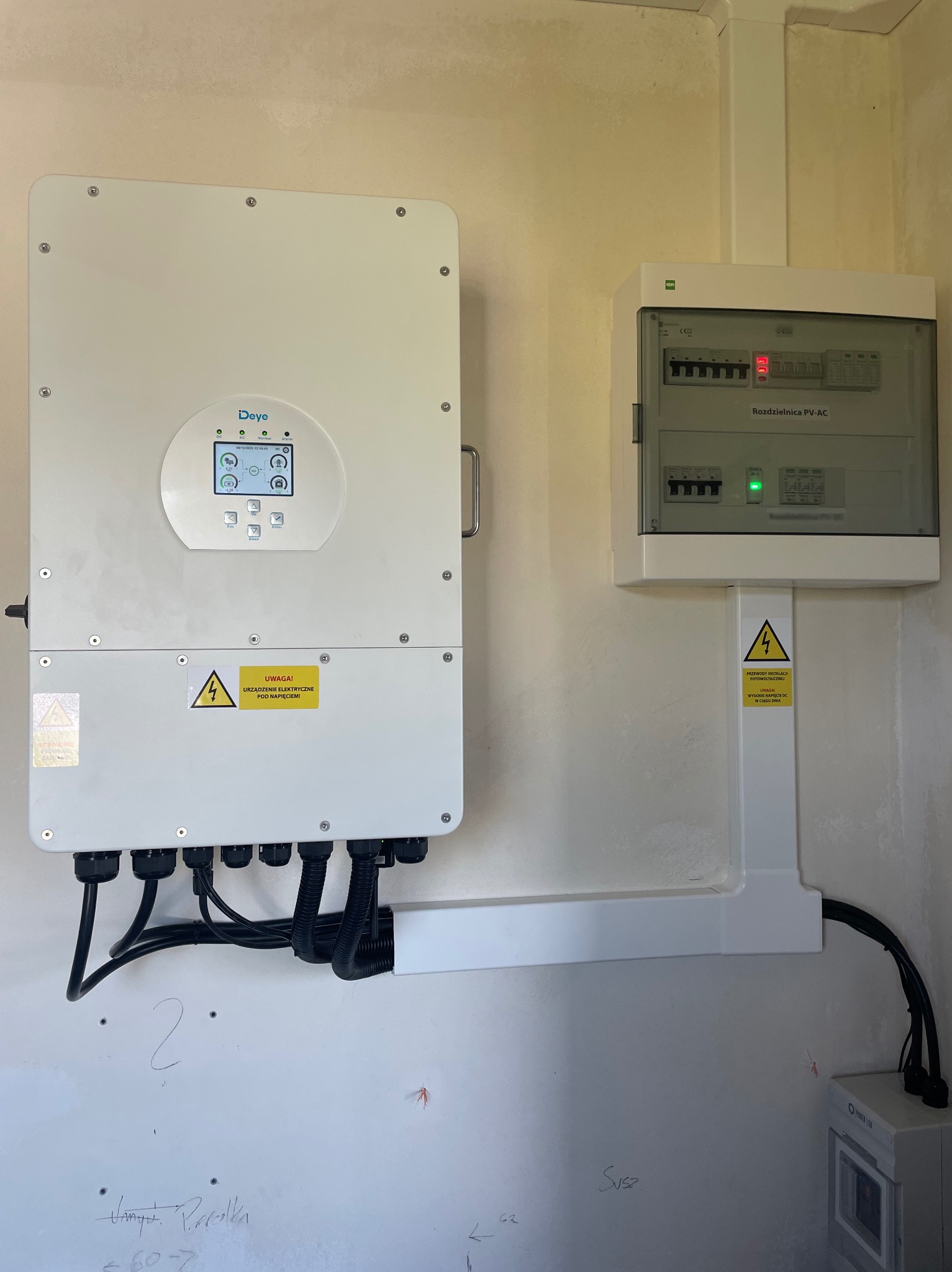 Biały inwerter Deye i rozdzielnica PV-AC na ścianie. Widoczne kable w peszlach. Żółte naklejki ostrzegawcze o napięciu. Montaż instalacji fotowoltaicznej wewnątrz budynku.