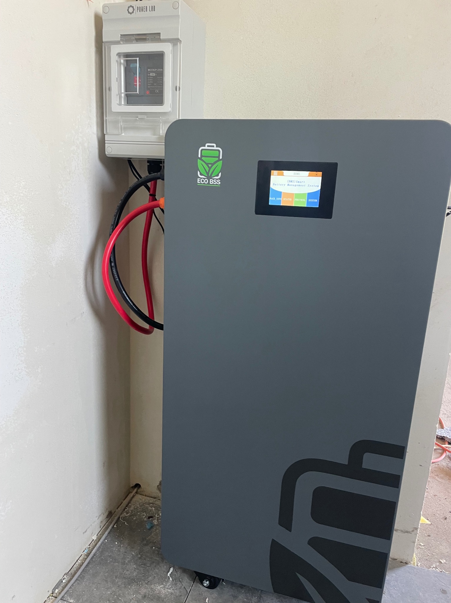 Szary magazyn energii ECO BSS z wyświetlaczem i podłączonymi kablami, obok biała skrzynka POWER LAD na ścianie. Widoczne kable i elementy instalacji w pomieszczeniu.