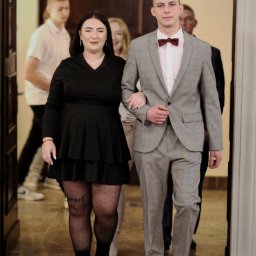 Fotografia Szymon Fraszewski - Para elegancko ubrana, mężczyzna w szarym garniturze i bordowej muszce, kobieta w czarnej sukience i rajstopach w kropki z napisem 'angel', w tle inne osoby wychodzące z pomieszczenia.