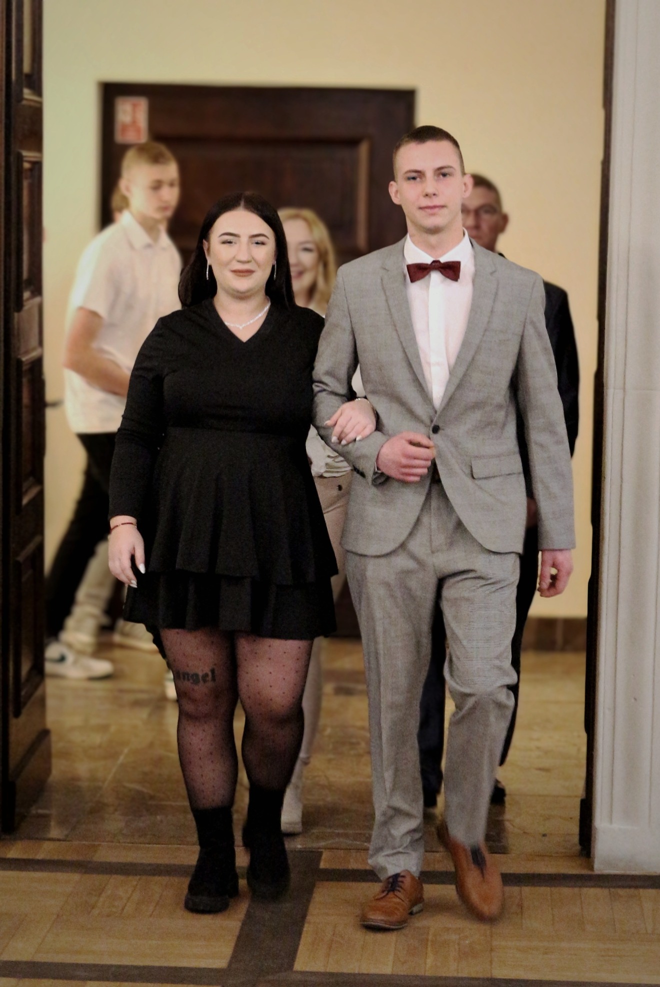 Para elegancko ubrana, mężczyzna w szarym garniturze i bordowej muszce, kobieta w czarnej sukience i rajstopach w kropki z napisem 'angel', w tle inne osoby wychodzące z pomieszczenia.