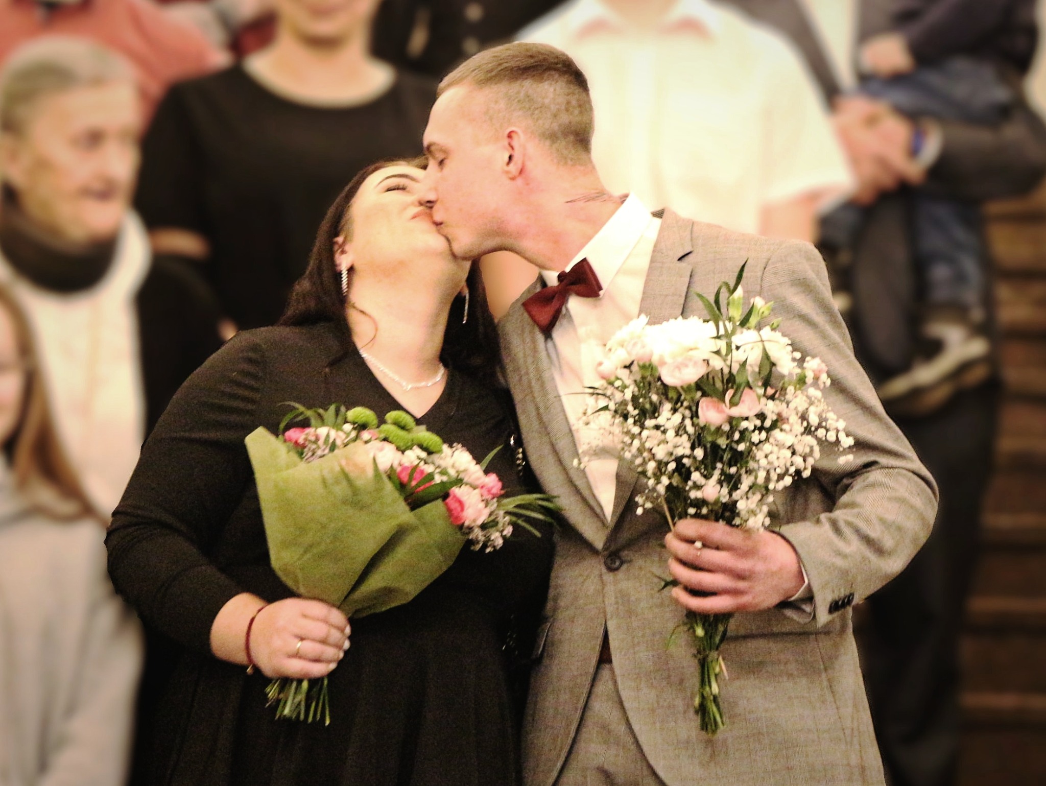 Para młoda w trakcie pocałunku, panna młoda w czarnej sukience trzyma bukiet w zielonym papierze, pan młody w szarym garniturze z bordową muchą trzyma bukiet z białych kwiatów, w tle goście.