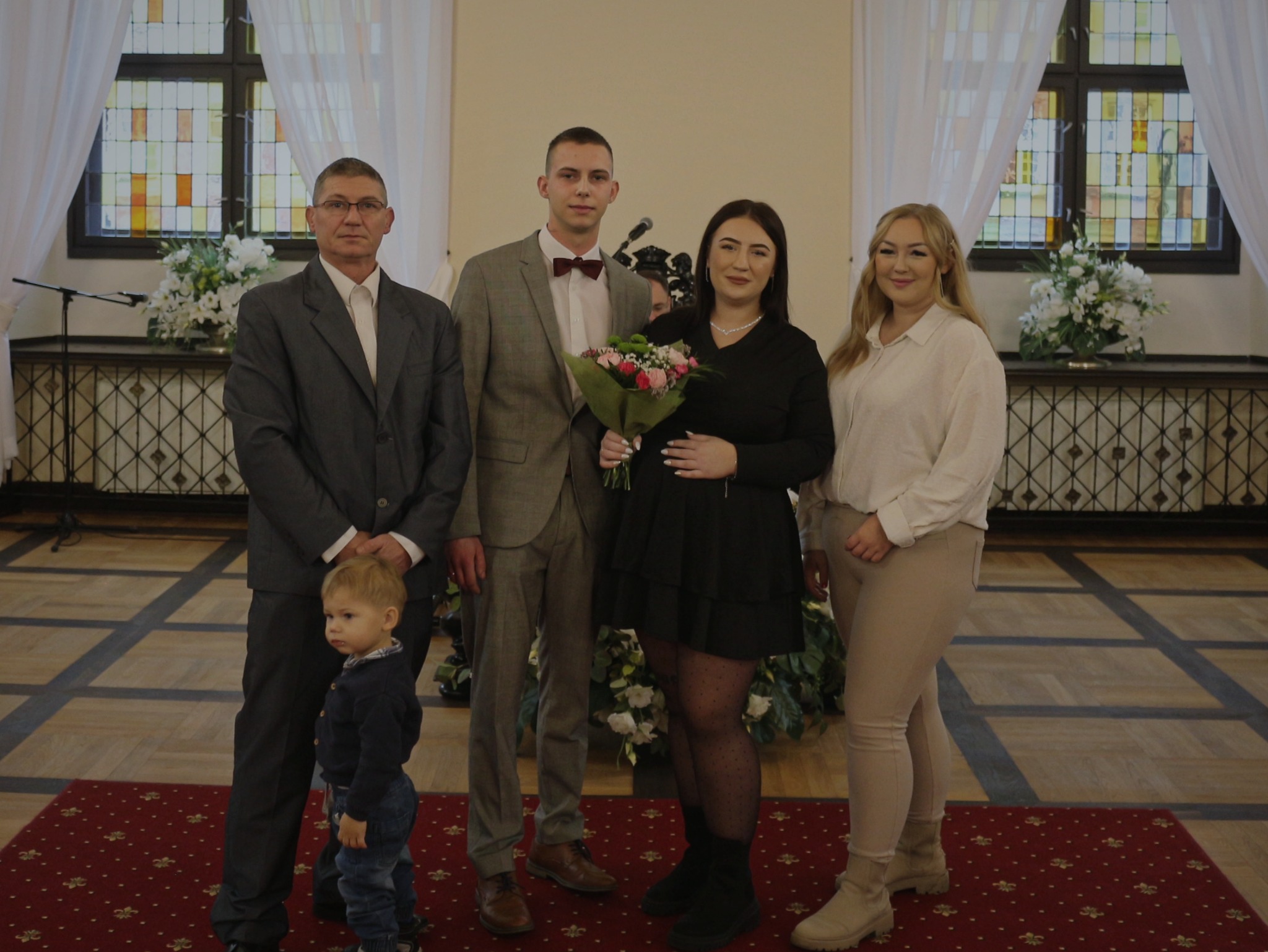 Grupa osób w eleganckich strojach pozuje do zdjęcia w przestronnym wnętrzu z dużymi oknami i kwiatowymi dekoracjami. Kobieta w czarnej sukience trzyma bukiet kwiatów, a obok niej stoi mężczyzna...