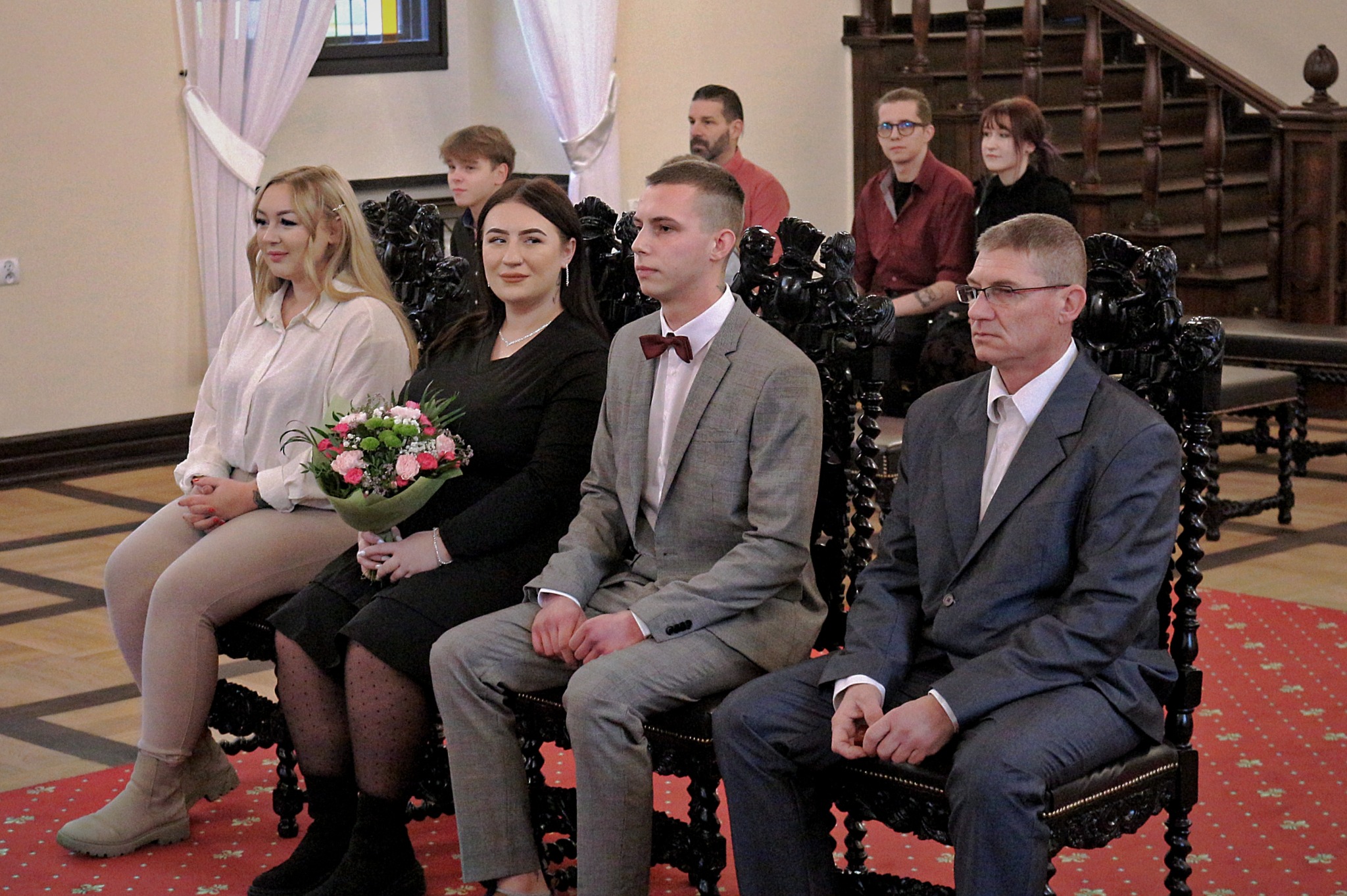 Goście weselni siedzą na ozdobnych krzesłach w kościele, czekając na rozpoczęcie ceremonii. Panna młoda trzyma bukiet kwiatów.