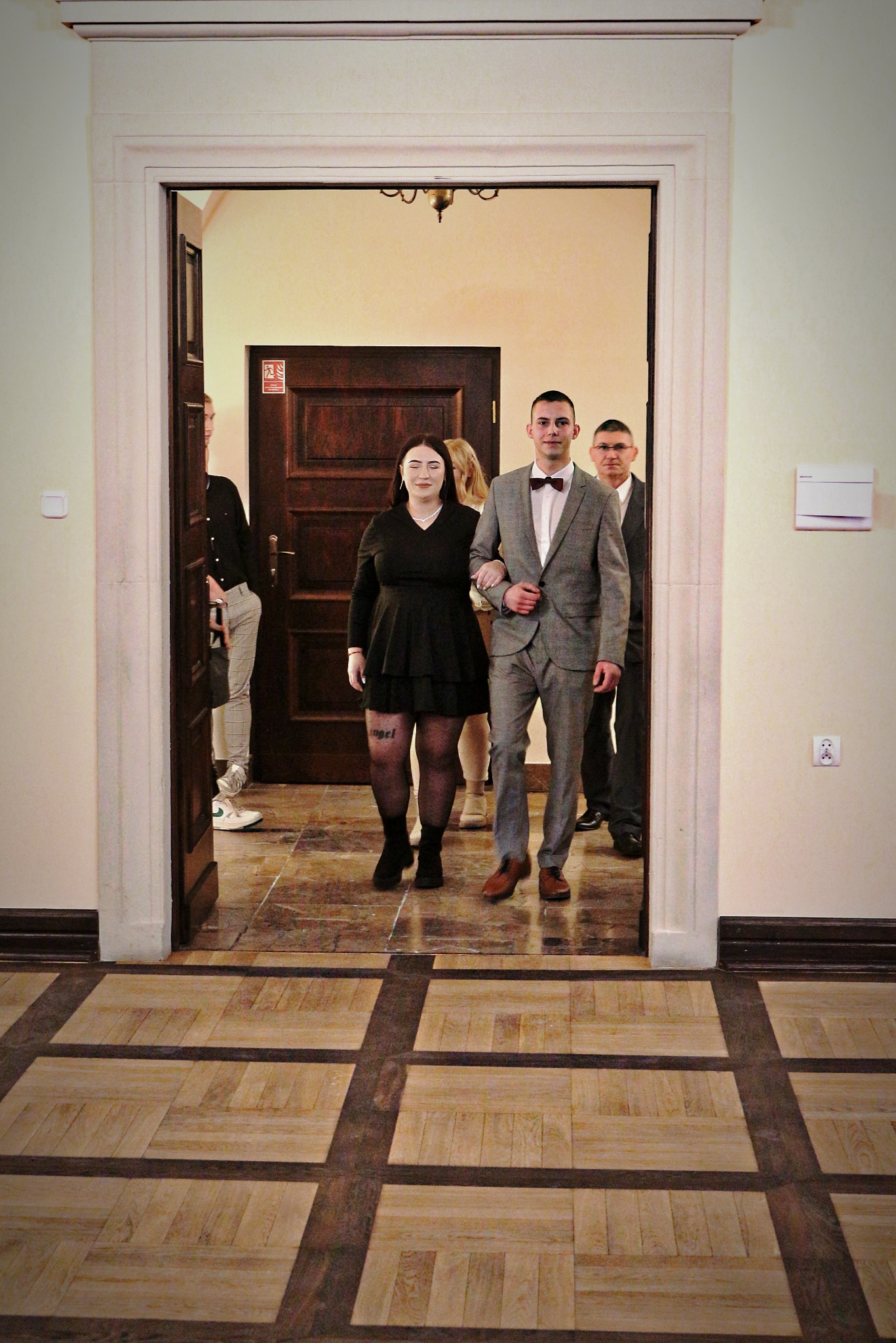 Elegancko ubrana para wchodzi do sali przez drzwi, za nimi widoczni inni goście, podłoga w geometryczny wzór.