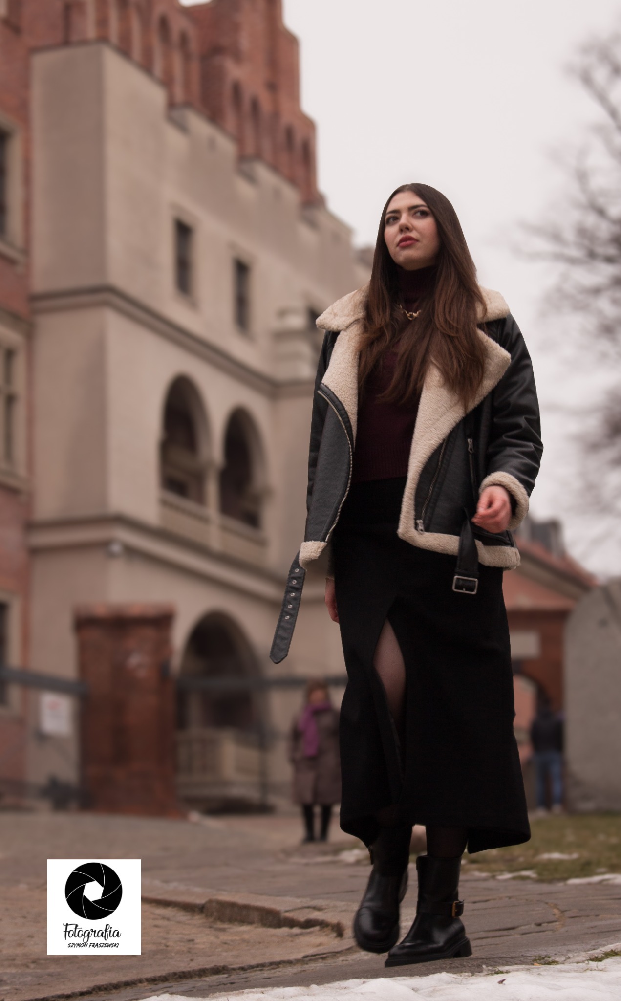 Kobieta w czarnej kurtce z futrzanym kołnierzem i długiej spódnicy z rozcięciem, pozująca na tle miejskiej architektury z cegły.
