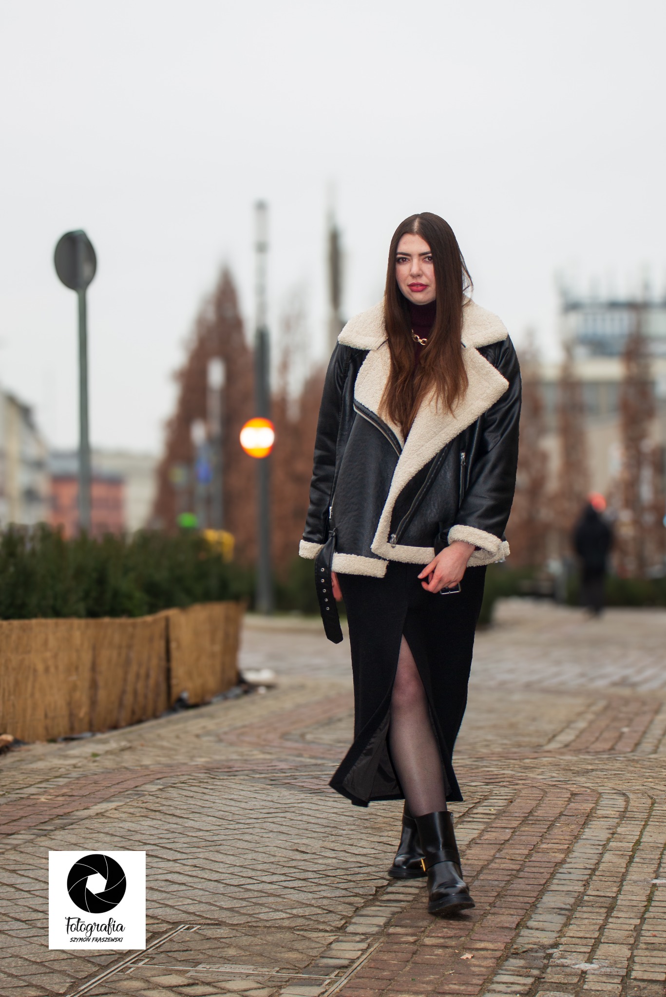 Kobieta w czarnej kurtce z futrzanym kołnierzem i długiej czarnej spódnicy z rozcięciem, pozująca na brukowanej ulicy w miejskiej scenerii.