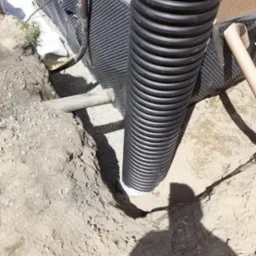 Czarna, karbowana rura pionowa w wykopie, obok ściany fundamentowej z izolacją kubełkową i drenażem obsypanym żwirem.