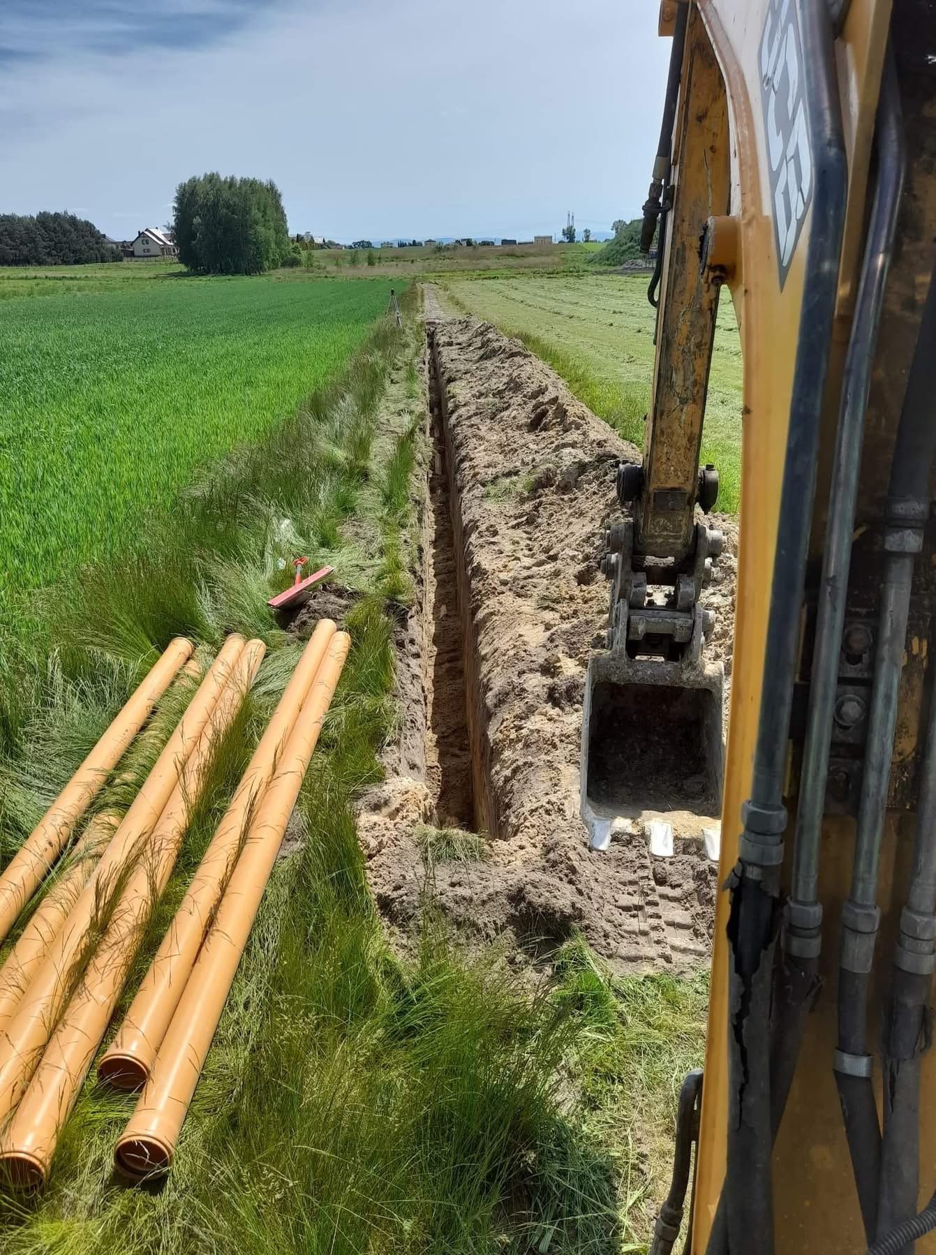 Żółta minikoparka częściowo widoczna z prawej strony kadru wykonuje wąski wykop w gruncie, w tle pole i budynki, obok wykopu leżą rury kanalizacyjne.
