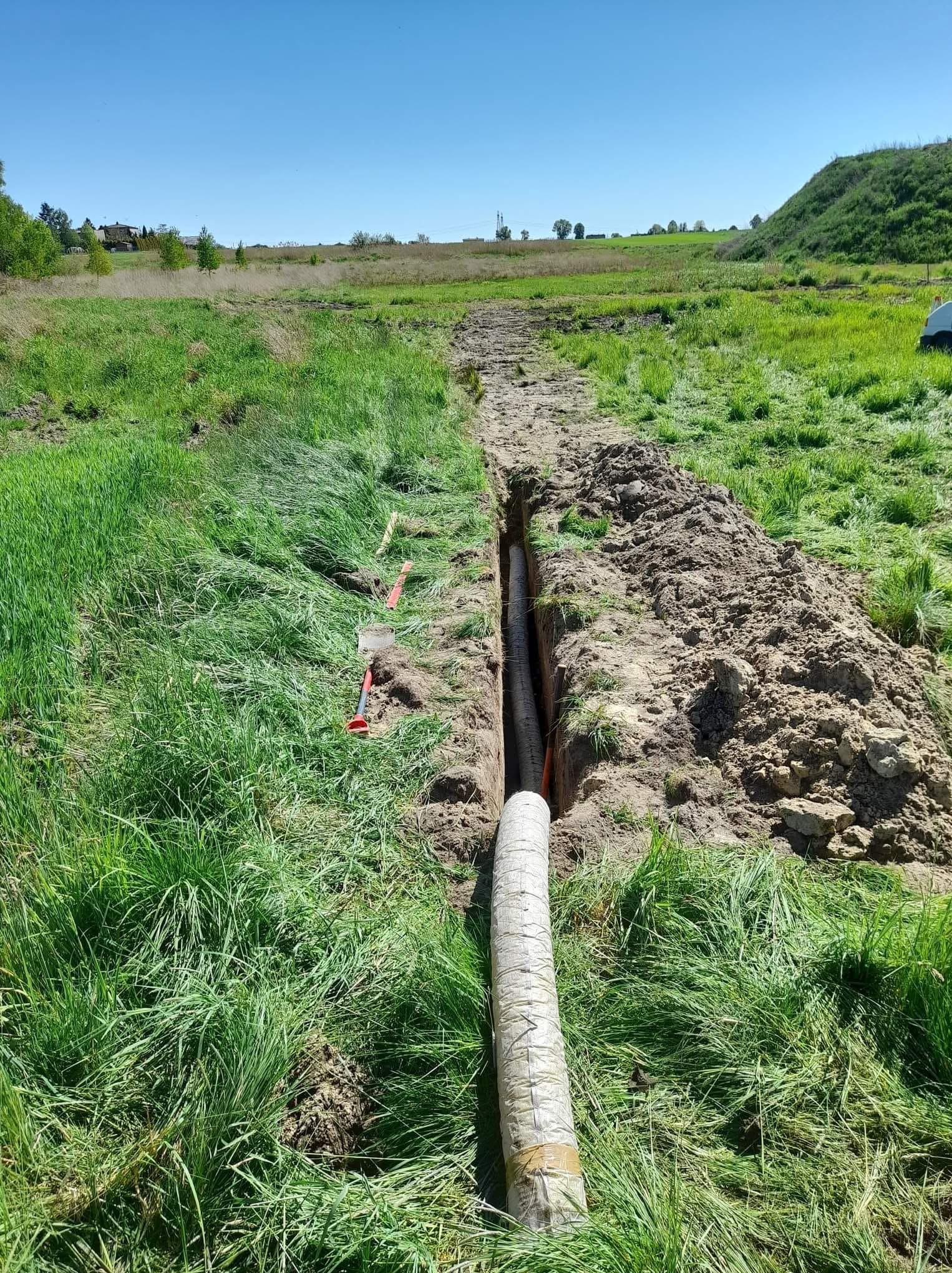 Rów w trawiastym terenie z częściowo zakopaną rurą w osłonie; widoczne narzędzia i wykopana ziemia